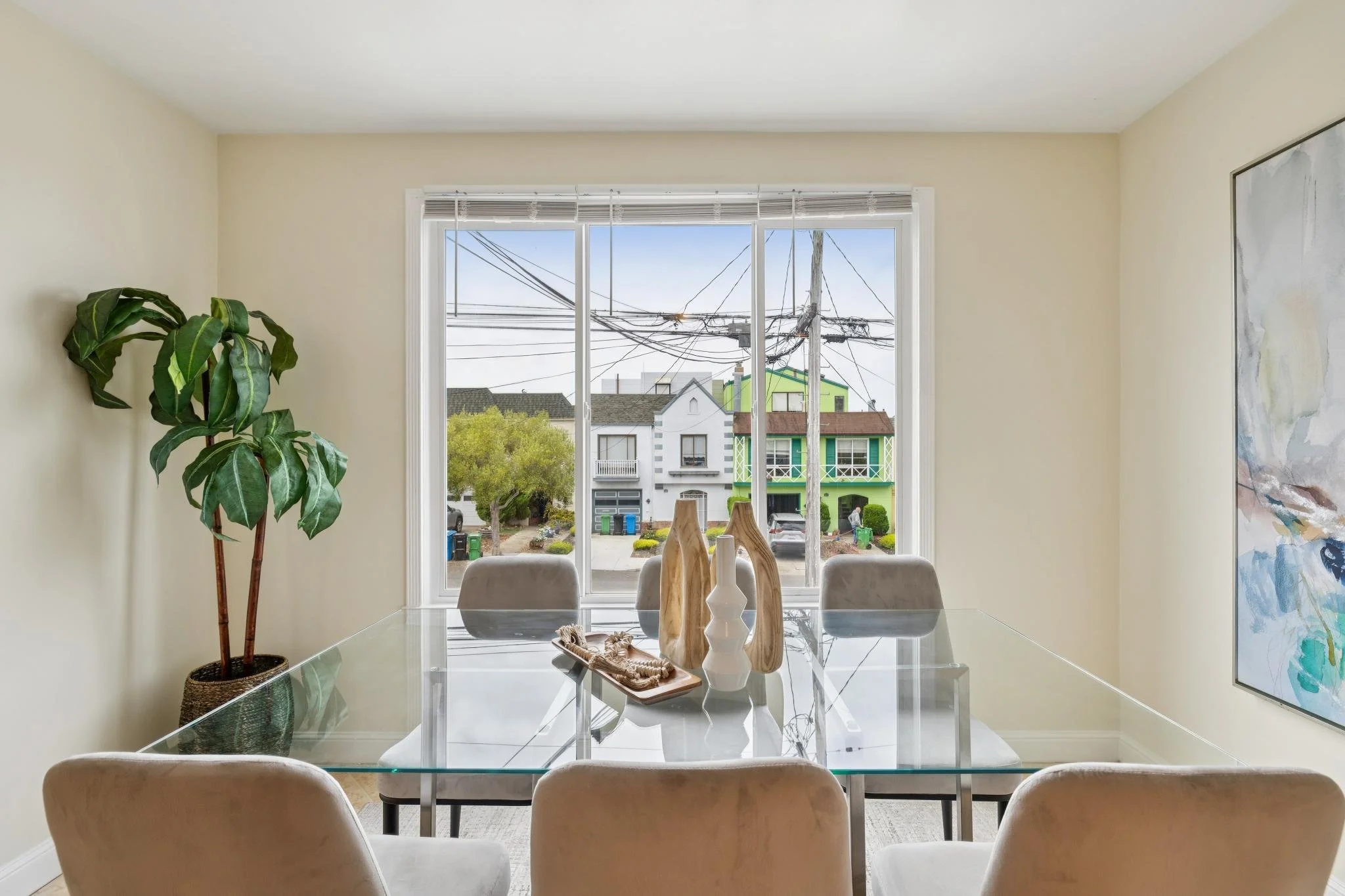 A dining room with a glass table, six beige chairs, a large window showing a neighborhood with a green house, trees, and utility poles, and a plant in a basket on the left.