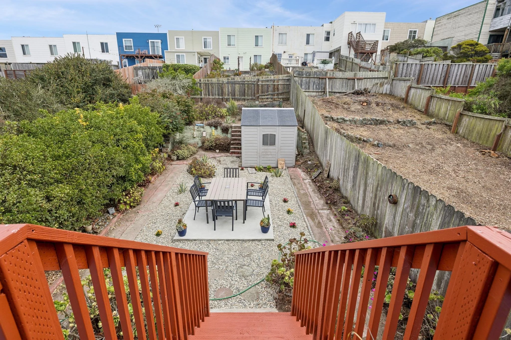 View from a balcony overlooking a backyard with a dining table, chairs, potted plants, a shed, and a sloped yard with a wooden fence; in the background, upper story houses with different colors are visible.