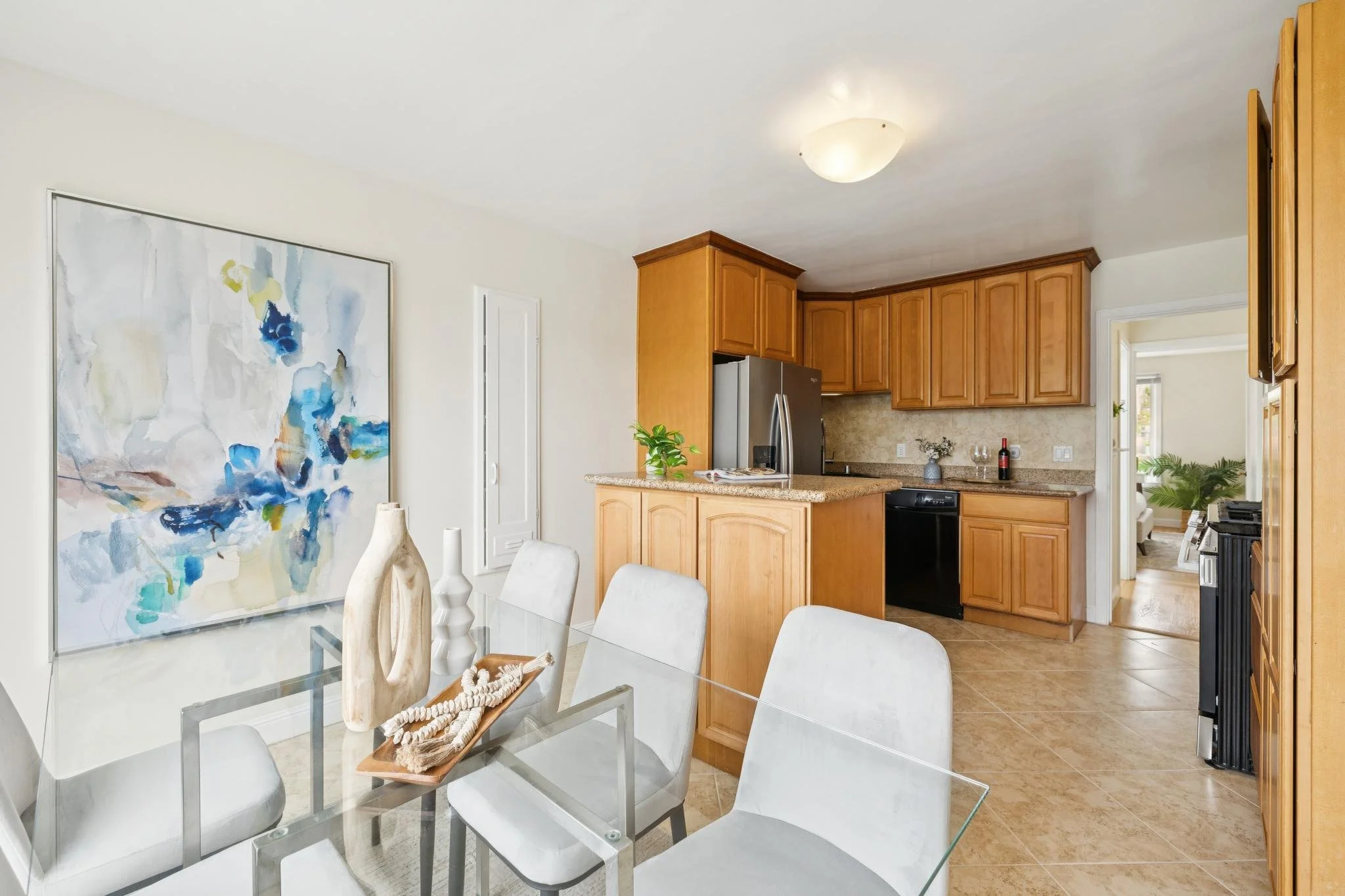 Interior view of a dining area connected to a kitchen with wooden cabinets, stainless steel refrigerator, and a granite countertop. A large abstract painting decorates the wall, and a glass-top dining table with white chairs and decorative vases is i