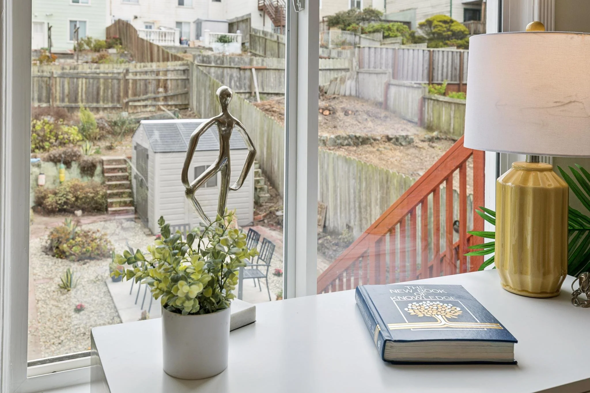 A windowsill with a small potted plant, a yellow lamp, and a book titled 'The New World Knowledge'. Outside the window, there is a backyard with a patio, garden, and old wooden fencing.