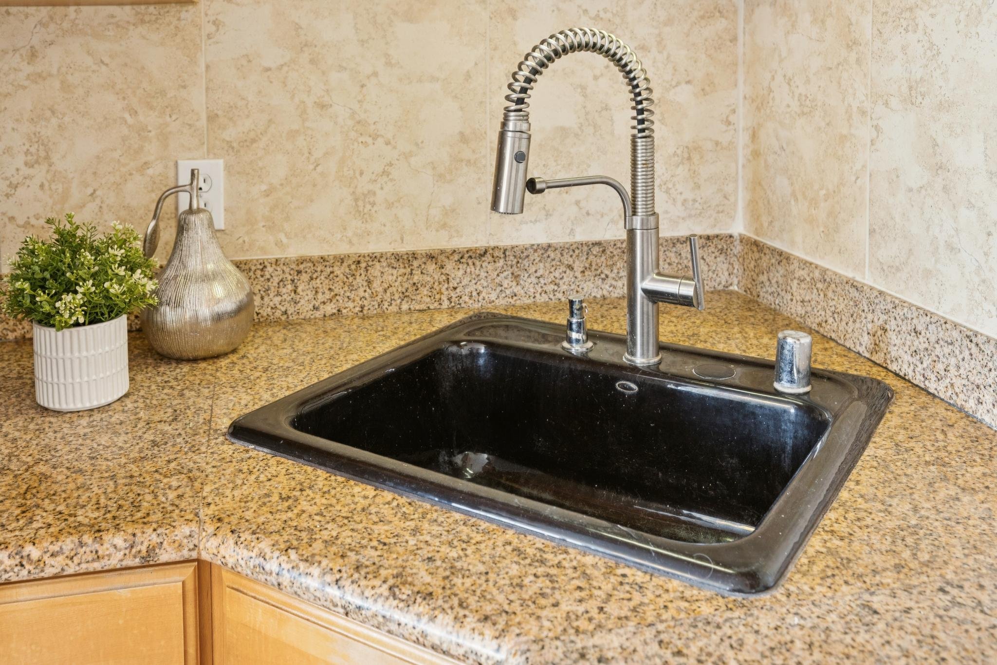 A kitchen sink with a modern metal faucet on a beige granite countertop. To the left of the sink, there is a small potted plant and a decorative silver-colored bottle. The background features beige tiled walls and wooden cabinet drawers below the cou