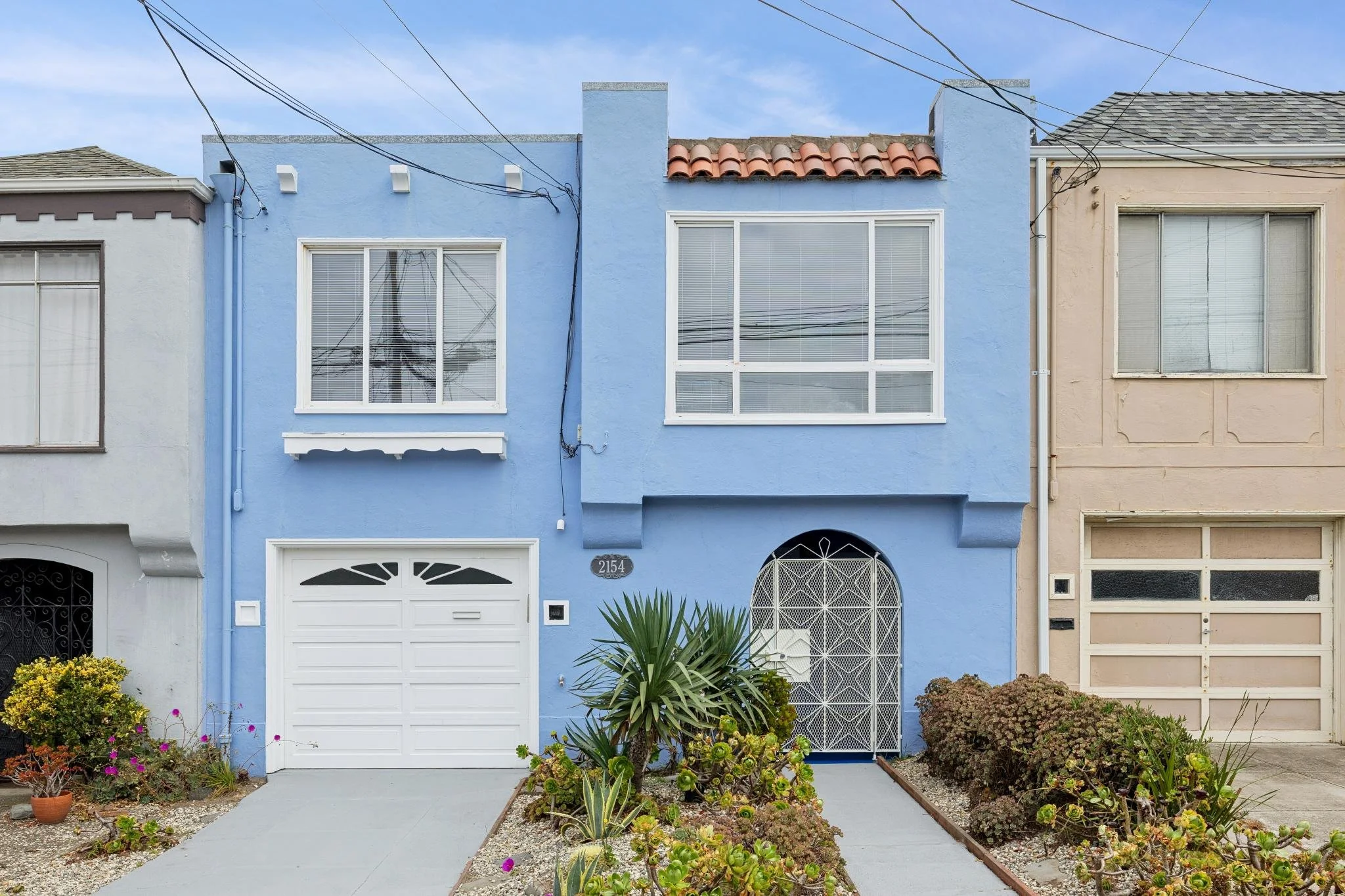 A row of colorful houses with front gardens, including a blue house with white garage door and windows, and a beige house with large window, plants, and shrubs in front.