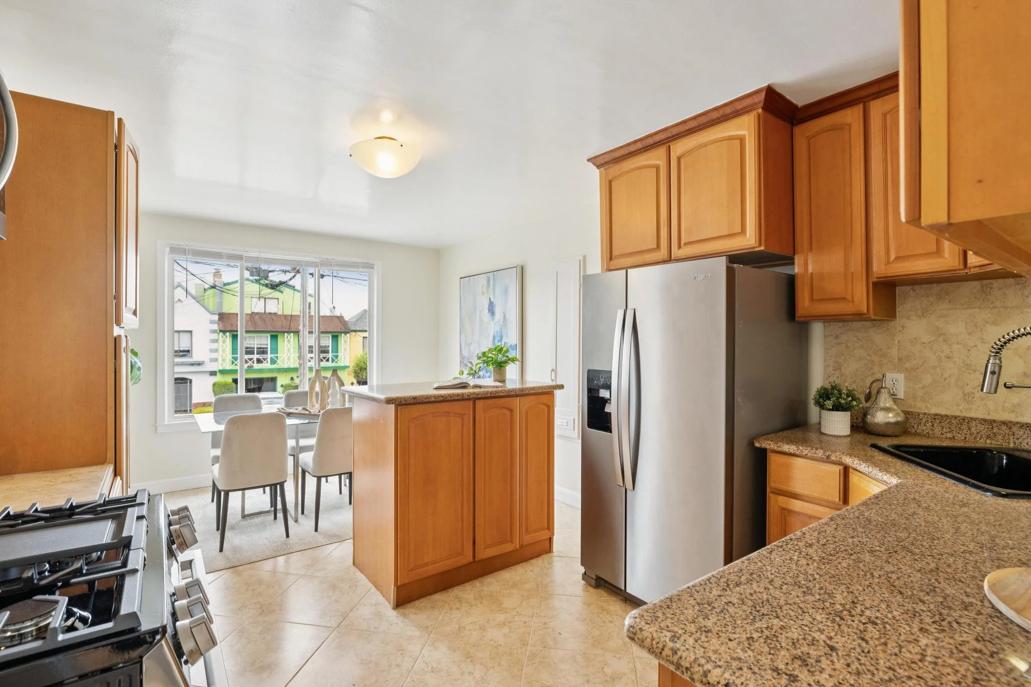 View of a bright kitchen with wooden cabinets, a stainless steel refrigerator, a granite countertop, a gas stove, a dining area with a glass table and white chairs, and a large window showing neighboring houses.