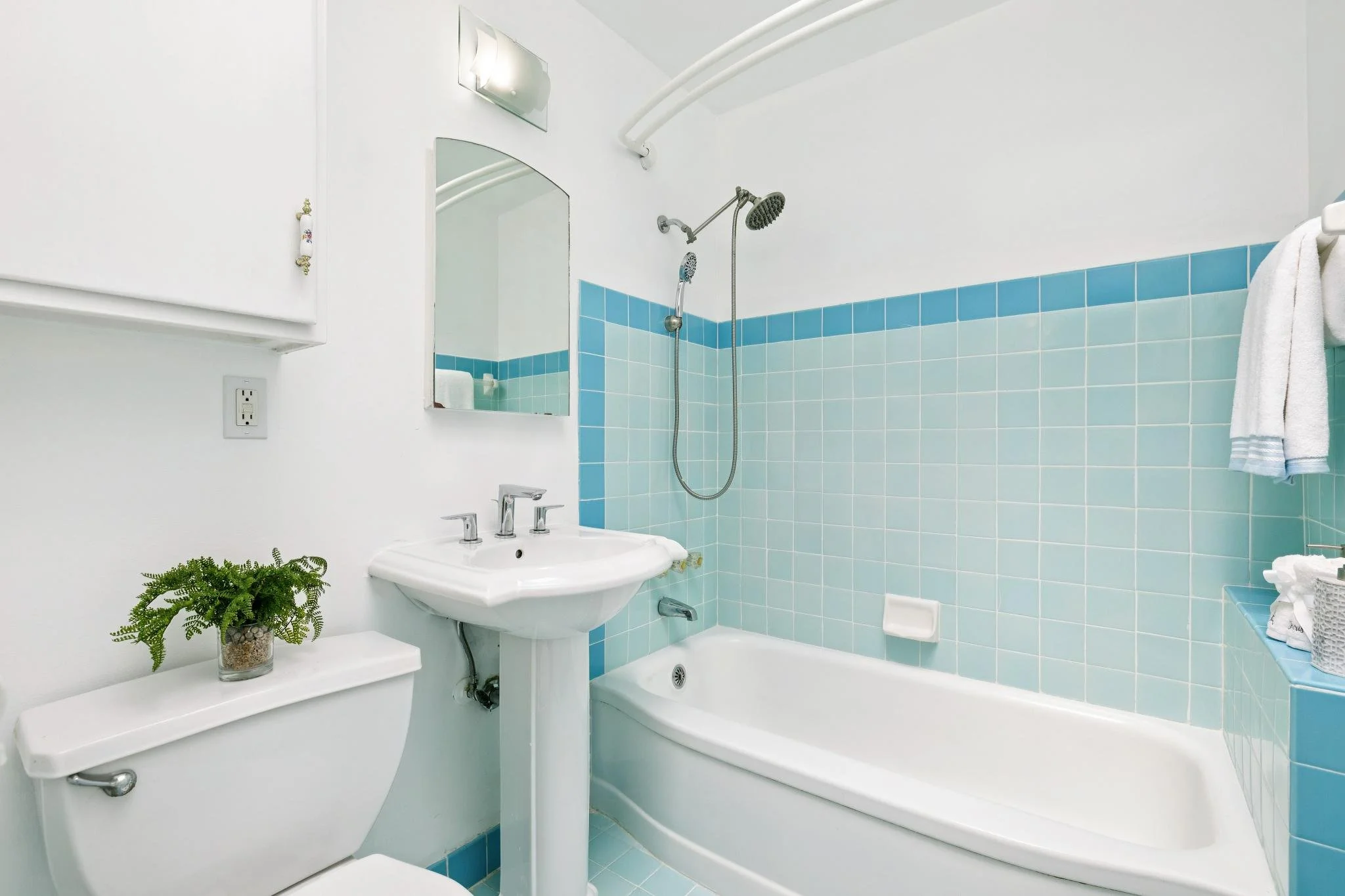 A bathroom with a white bathtub, blue tiled wall, white sink with silver faucet, mirror, white towel, potted plant, and light fixture.