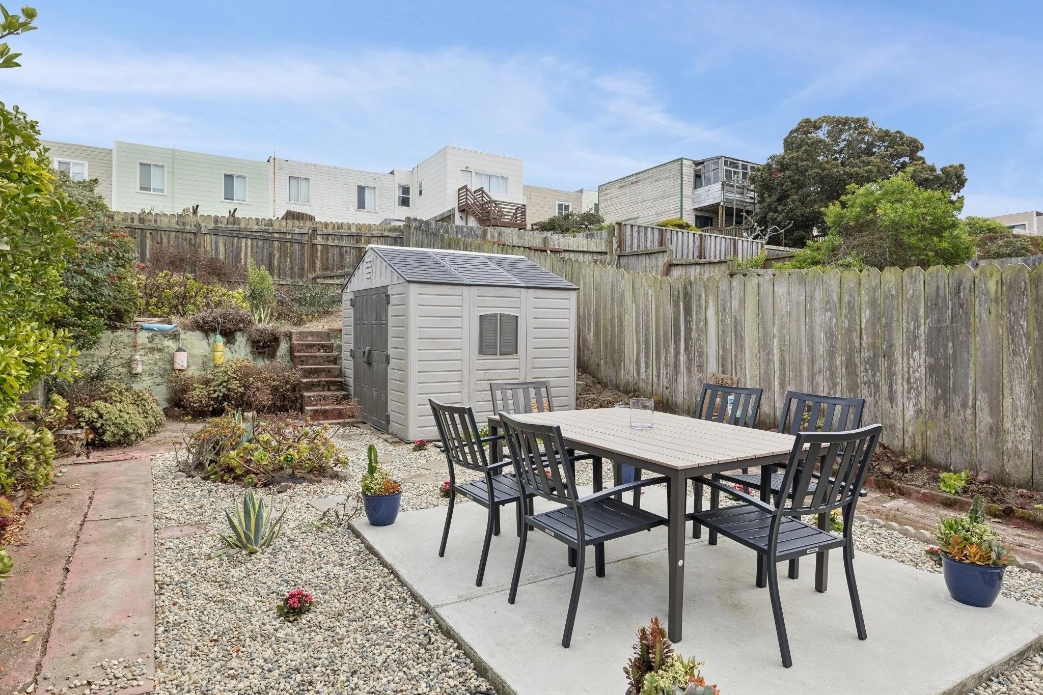 Backyard patio with black dining table and chairs, potted plants, and small garden beds, enclosed by weathered wooden fences, with a small storage shed, multicolored stones, and neighboring houses in the background under a blue sky.