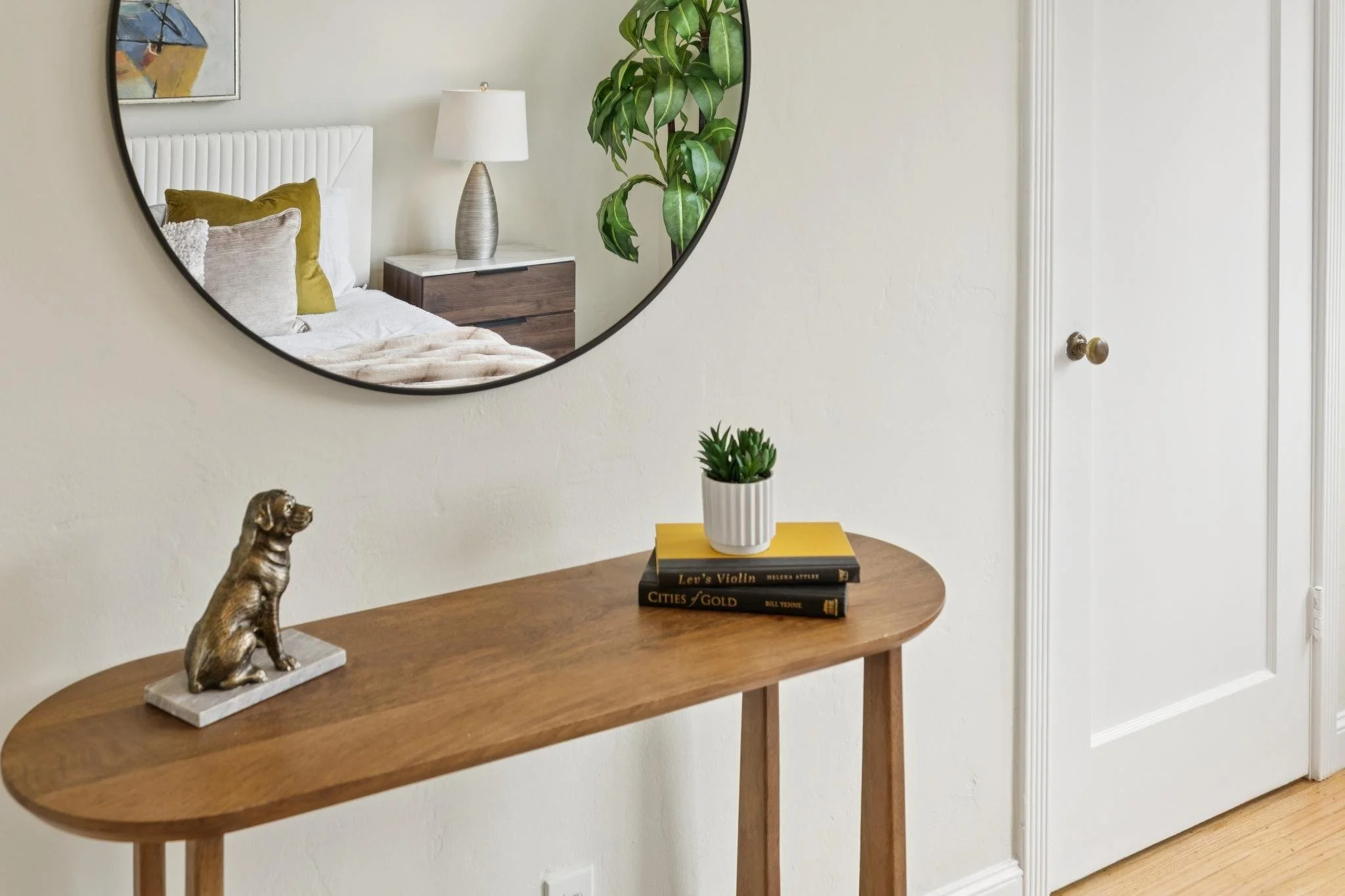 Interior view of a hallway with a wooden console table, a gold dog sculpture, a small potted plant, and a stack of three books. A large round mirror reflects a bedroom with a white bed, several pillows, a nightstand, a lamp, and a piece of artwork. A