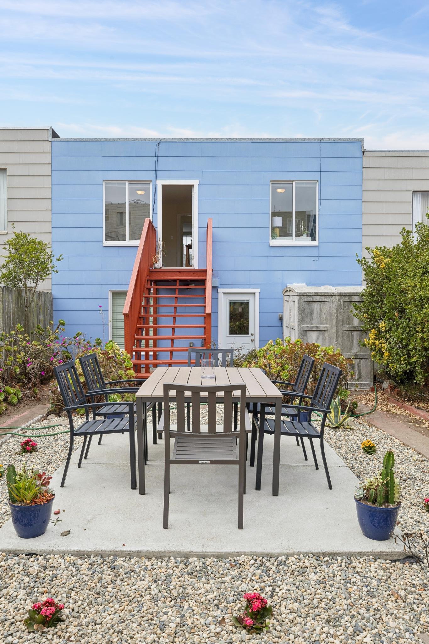 Backyard patio with a metal dining table and eight chairs, two large potted succulents, gravel ground, and a blue two-story house with a red staircase and a door and windows