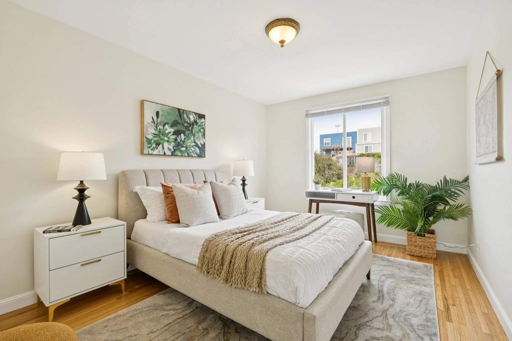 A bedroom with a beige upholstered bed, white bedding, and decorative pillows. There are two nightstands with lamps, one with books. A large window with white blinds shows neighboring houses. Artwork and a hanging fabric wall decoration are on the wh