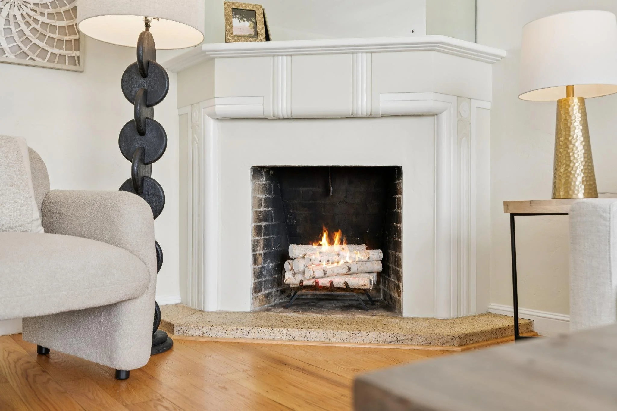 Living room with a white fireplace mantel, a fire burning inside, on the right a gold textured lamp, on the left a beige upholstered armchair, a black decorative chain sculpture next to it, and a beige carpeted floor.