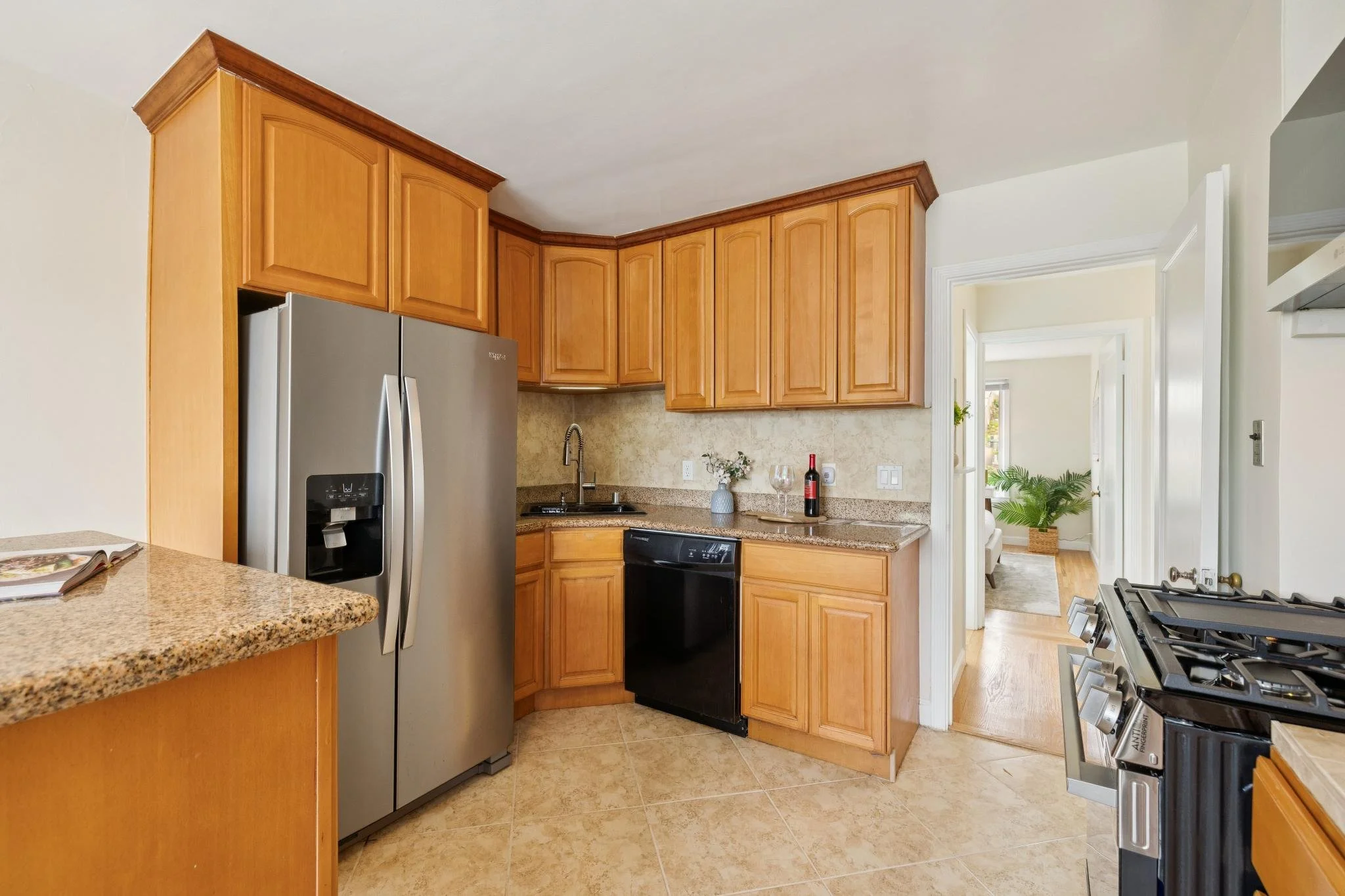A kitchen with wooden cabinets, a refrigerator, a granite countertop, a sink, and a stove, with a doorway leading to a bright room with wooden floors, a potted plant, and a chair.