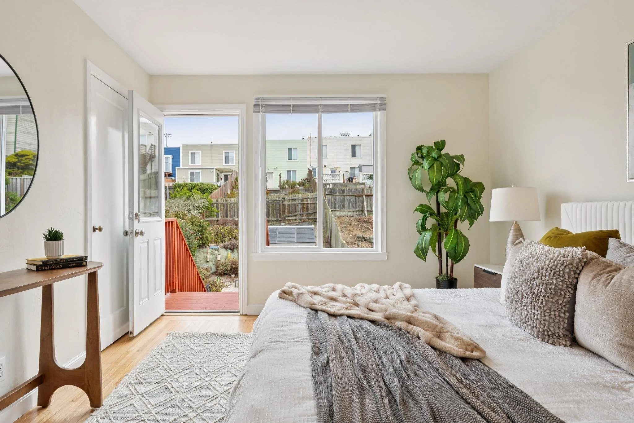 Bright living room with large window overlooking neighboring houses, beige walls, a potted plant, a table with books, and a bed with textured pillows.