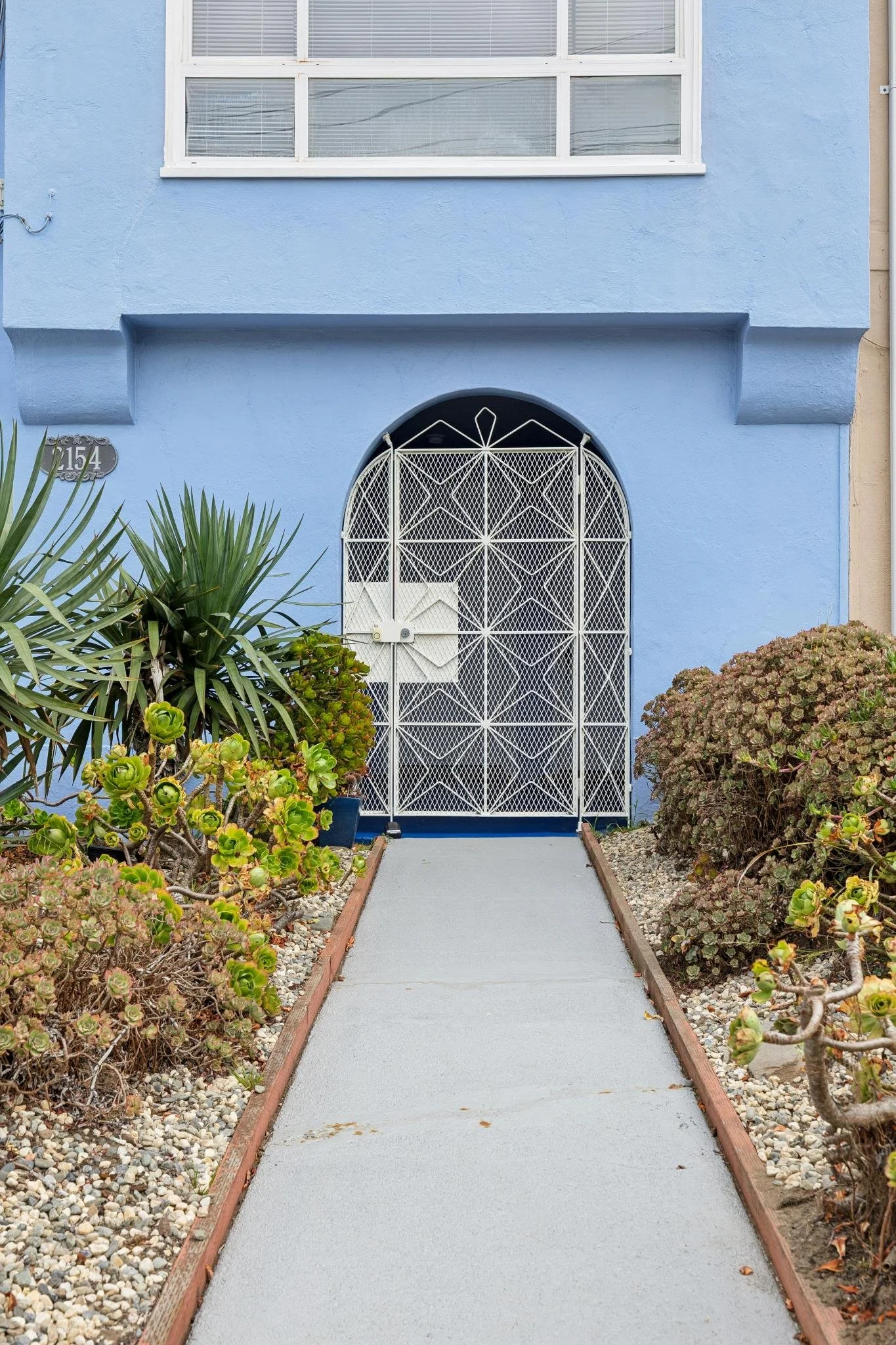A pathway leading to a blue house with a white decorative iron gate in front. There are green and brown bushes on both sides of the pathway, with a gravel border.