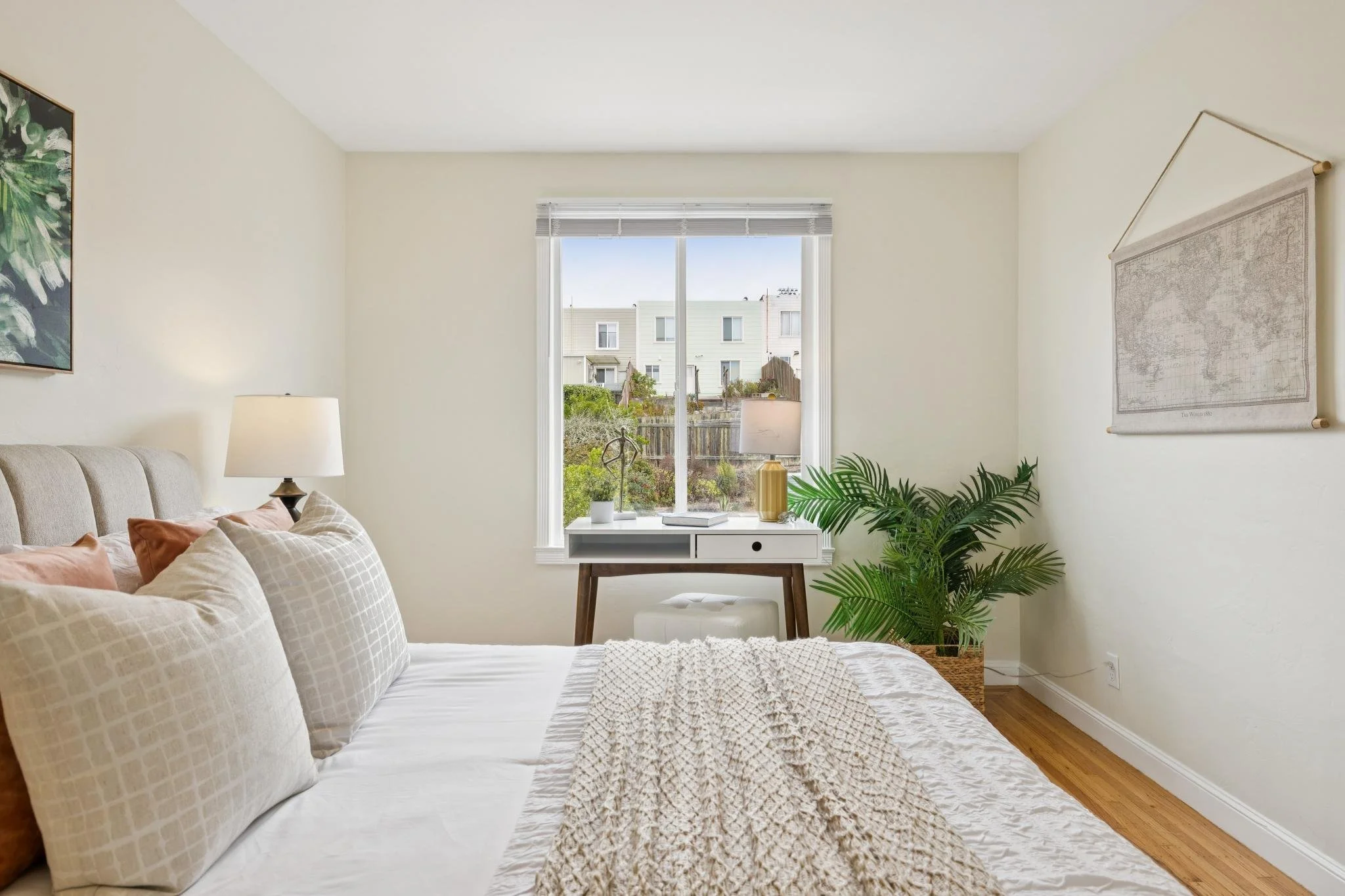 A cozy bedroom with beige walls and hardwood floors, featuring a bed with white and beige bedding, a nightstand with a lamp, a window with blinds showing neighboring houses, and various plants and wall art.