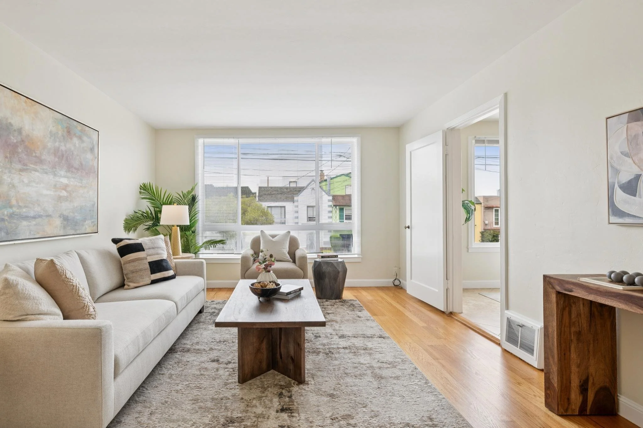 Bright living room with white sofa, armchair, wooden coffee table, large window with blinds, artwork on walls, and decor including potted plants and vases.