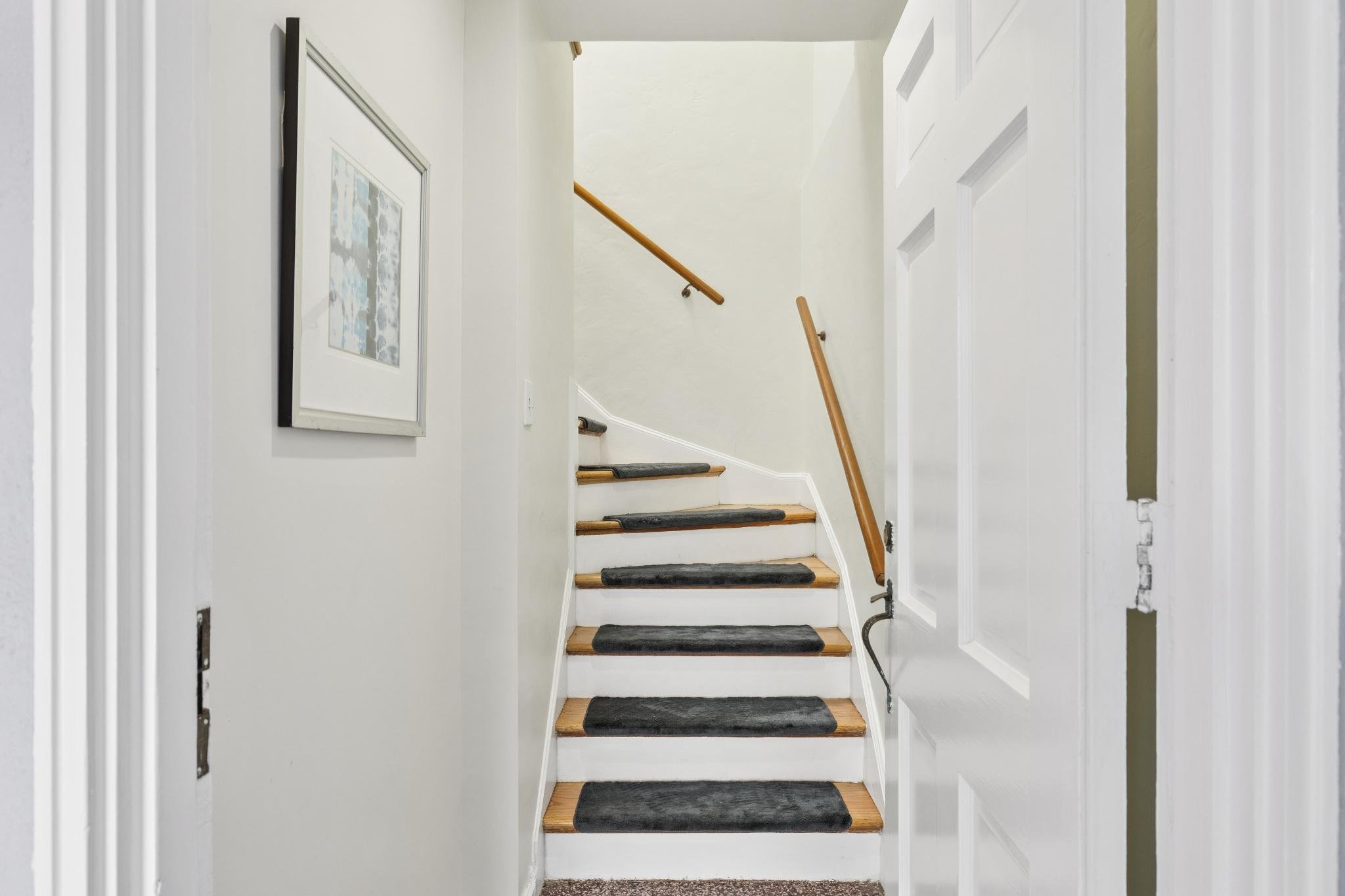 Indoor staircase with black carpeted steps, wooden handrails on both sides, white walls, and framed artwork on the left wall.