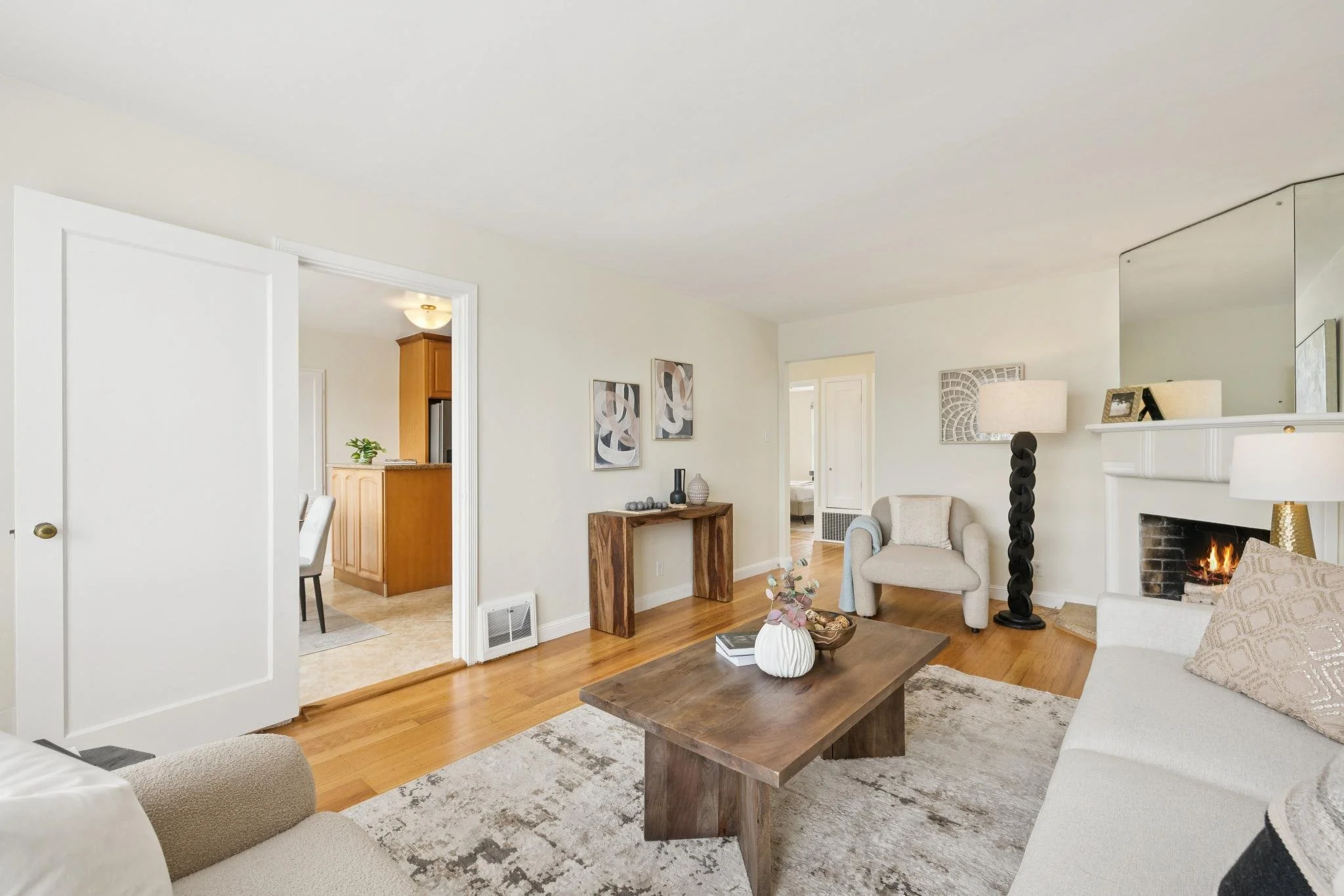 Living room with white walls, wooden floor, fireplace with lit fire, beige armchair, black sculpture lamp, wooden coffee table with vases, framed wall art, and adjacent kitchen area.