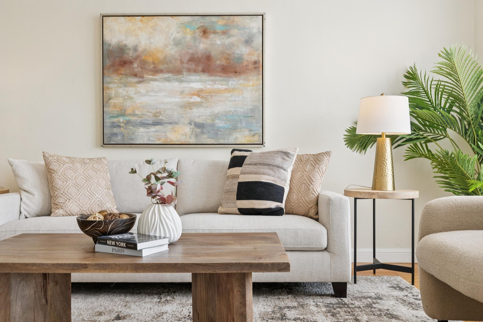 Living room with a beige sofa decorated with patterned pillows, a wooden coffee table with books and vases, a gold table lamp, a potted plant, and an abstract painting on white wall.