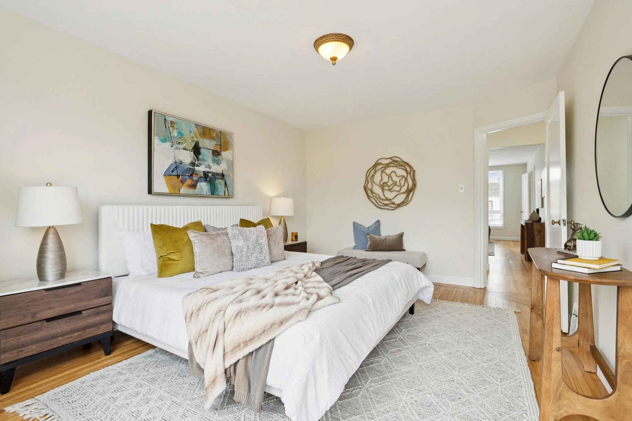 A cozy bedroom with a white bed, decorative pillows, and a faux fur throw. There are two lamps on nightstands, a colorful abstract painting above the bed, and a circular metal wall art piece. A wooden side table with books and a plant is near the doo