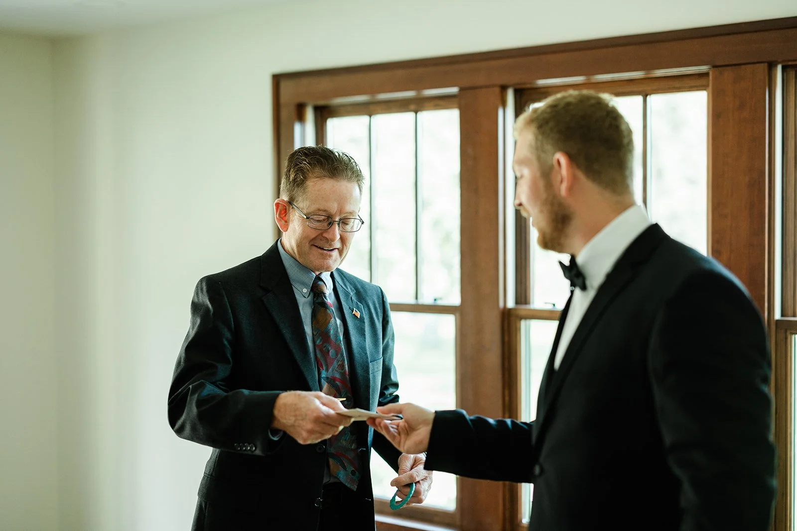 A man in a tuxedo with a bow tie handing a business card to another man in a suit with glasses and a patterned tie, indoors near a window.