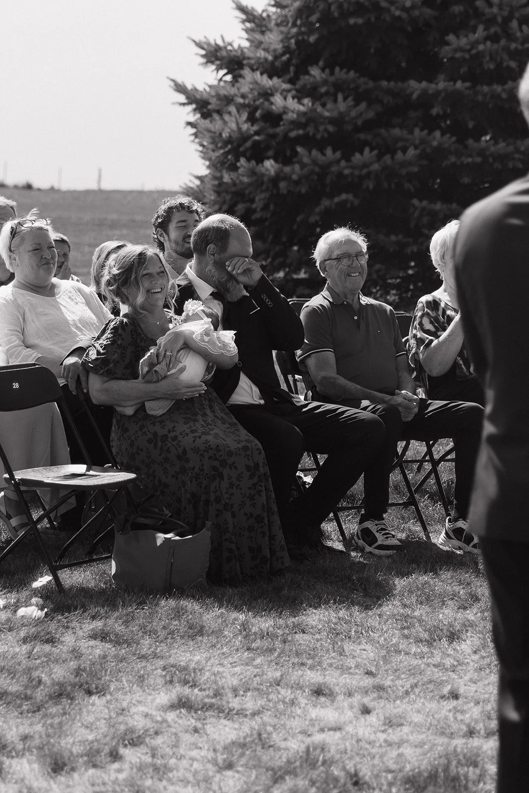 Black and white photo of a group of people sitting outdoors on chairs, smiling and laughing during a joyful event, with a large tree and open sky in the background.