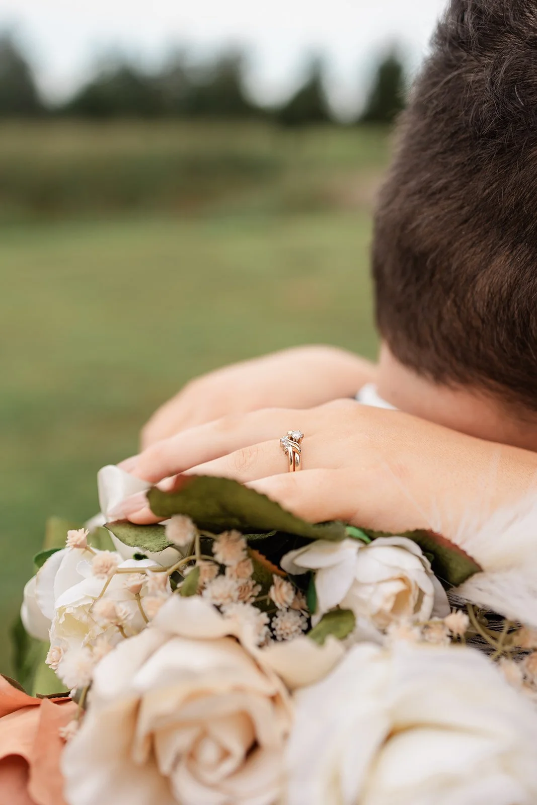 Close-up of a person resting on a bouquet of white roses and other flowers, with an engagement ring on their finger.