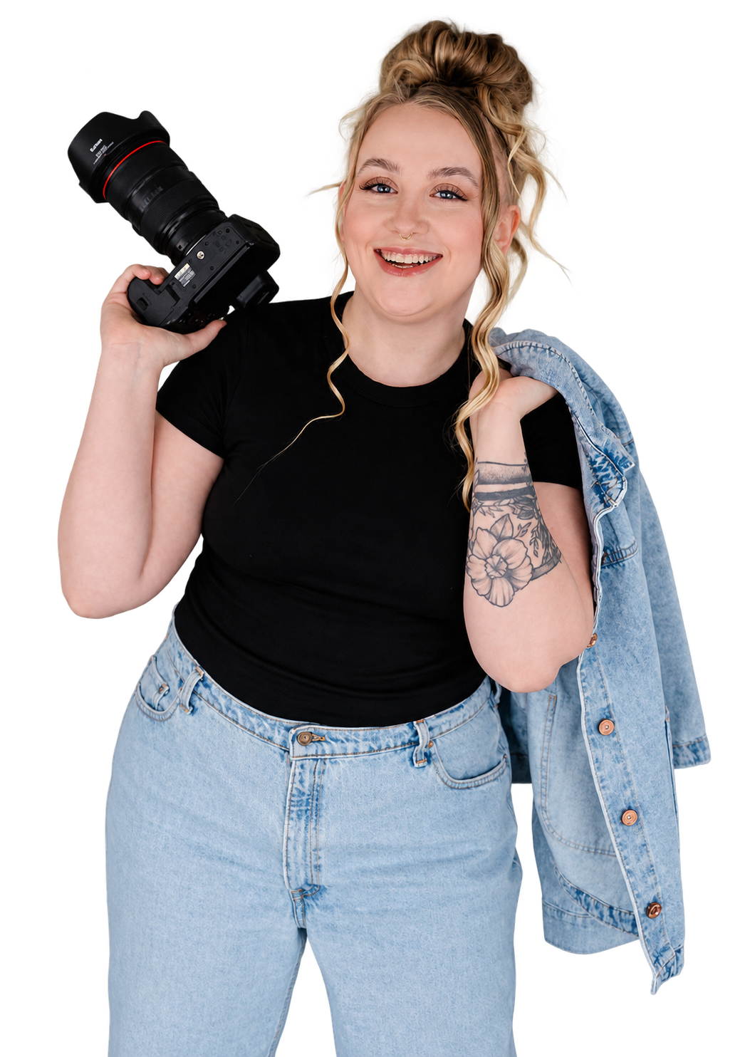 Young woman smiling, holding a camera over her shoulder, with a denim jacket draped over her shoulder and wearing a black t-shirt and light-wash jeans.