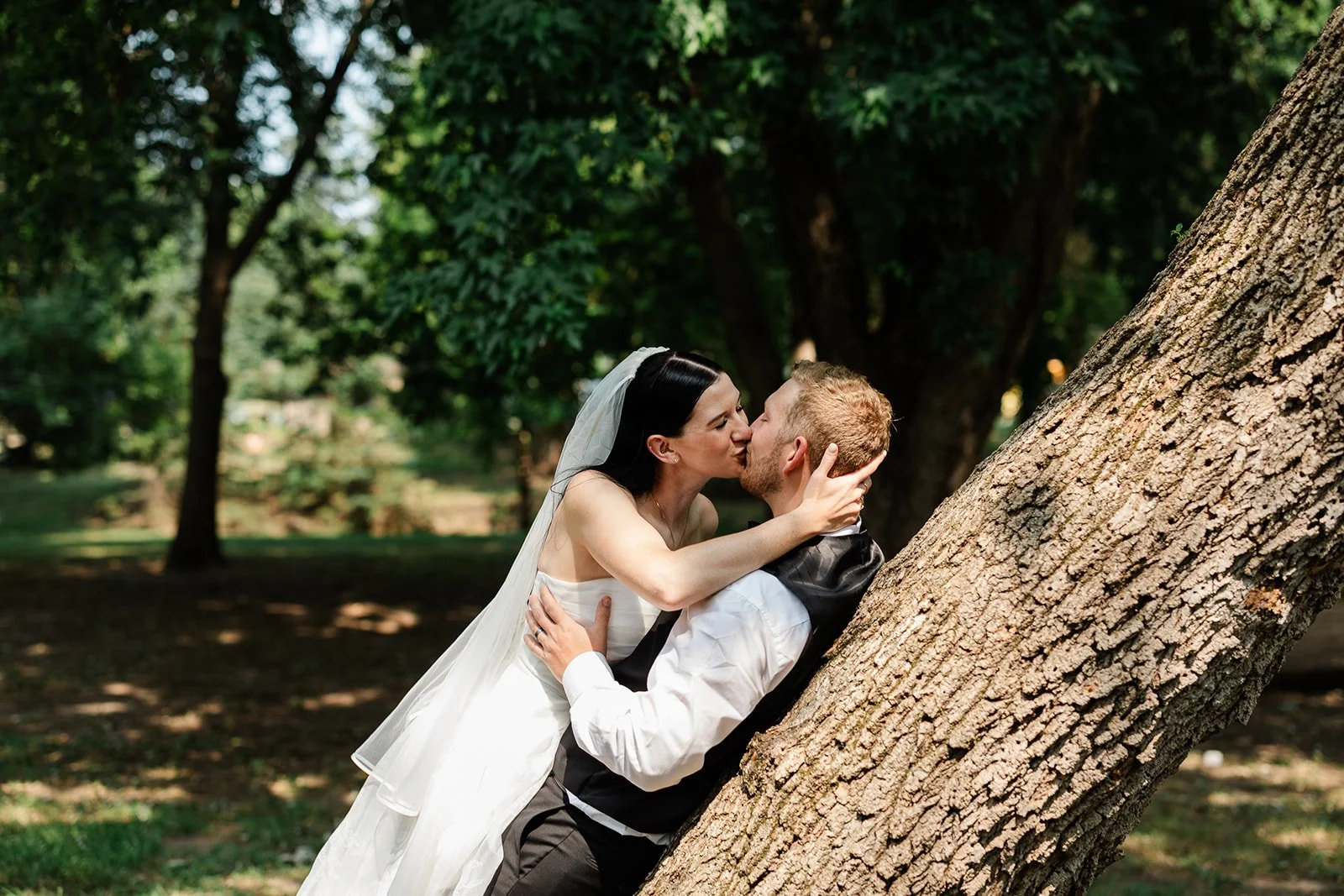 Bride and groom kissing outdoors under trees, with the bride in a white wedding dress and veil and the groom in a white shirt and black vest, leaning against a large tree trunk.