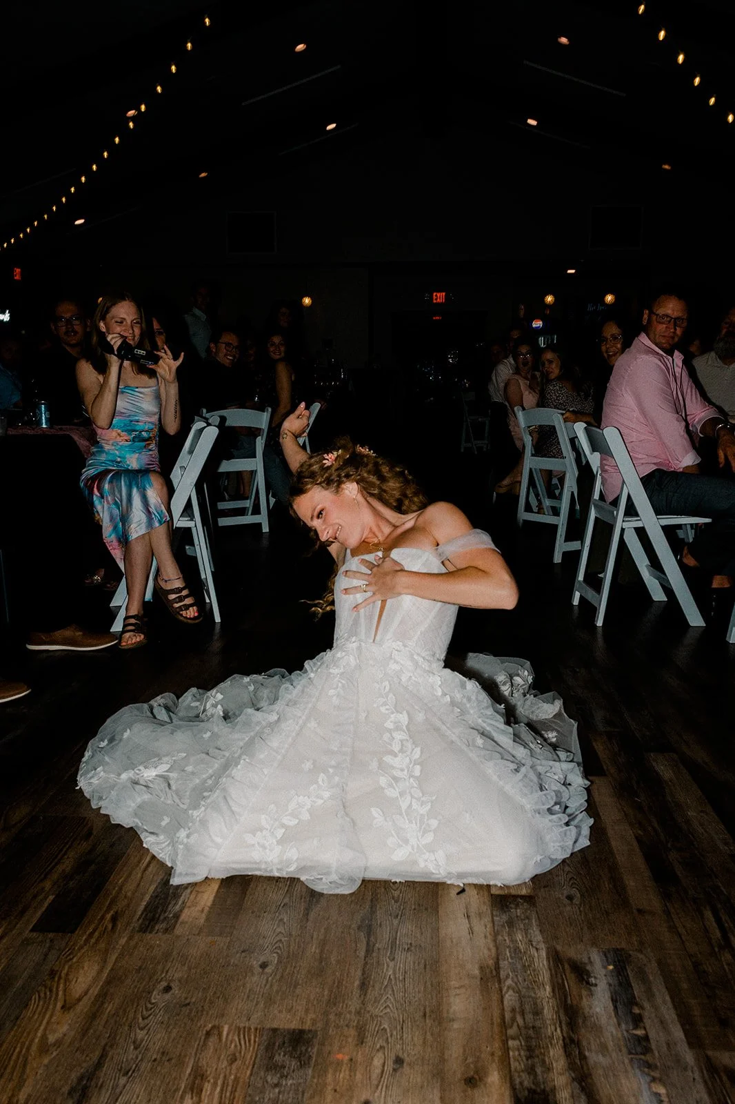 A bride in a white wedding dress sitting on the floor while dancing at her wedding reception with guests seated at tables in the background.