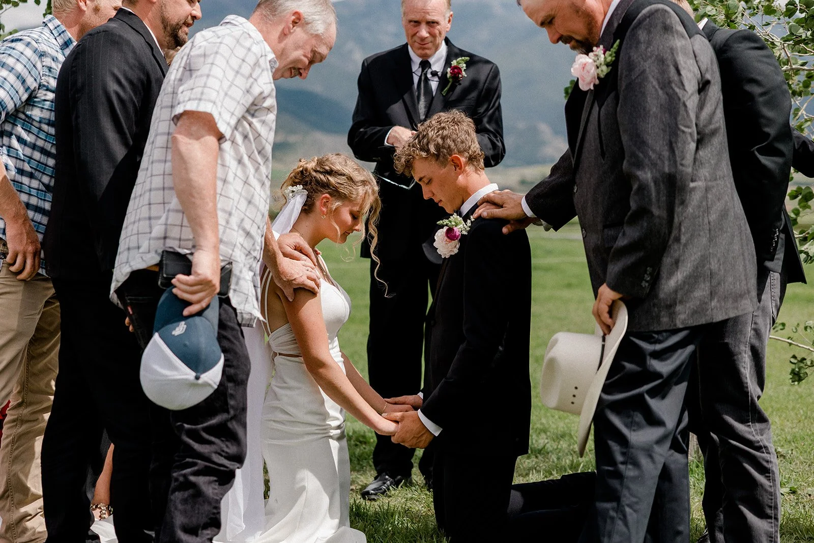 A wedding ceremony outdoors with a couple kneeling and holding hands, surrounded by family and friends in a grassy field under cloudy skies.