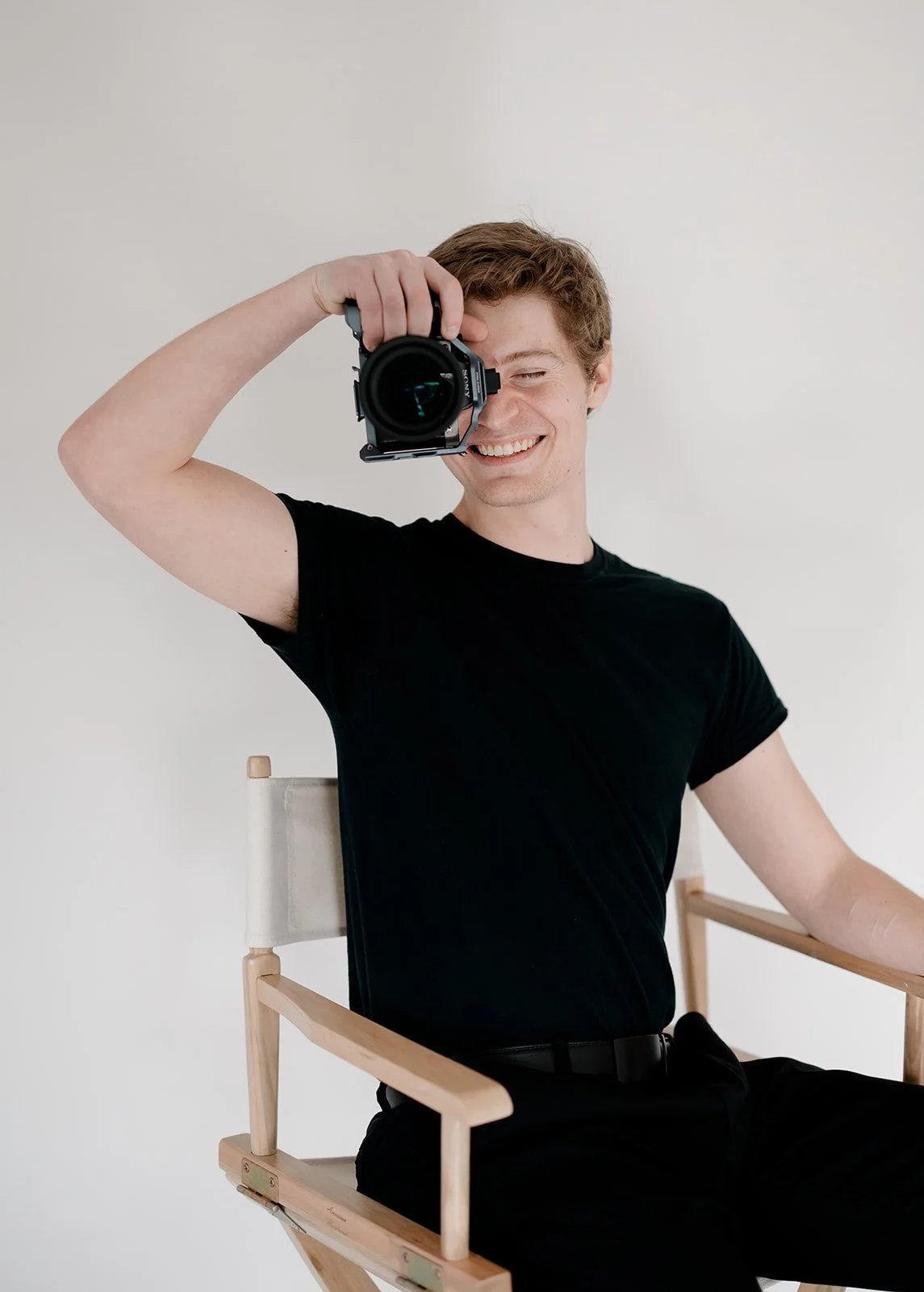 Smiling young man sitting on a director's chair, holding a camera, against a plain white background.