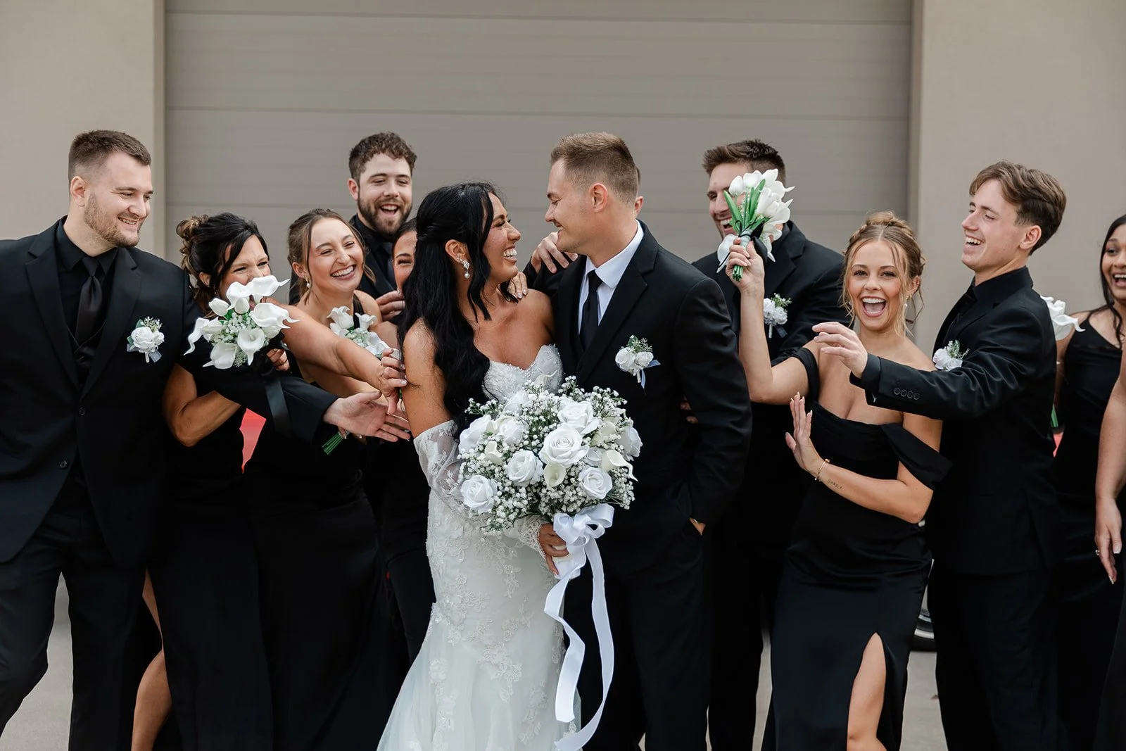 A wedding party of ten people, including the bride and groom, celebrating and smiling with flowers and formal attire, standing outside near a large gray door.