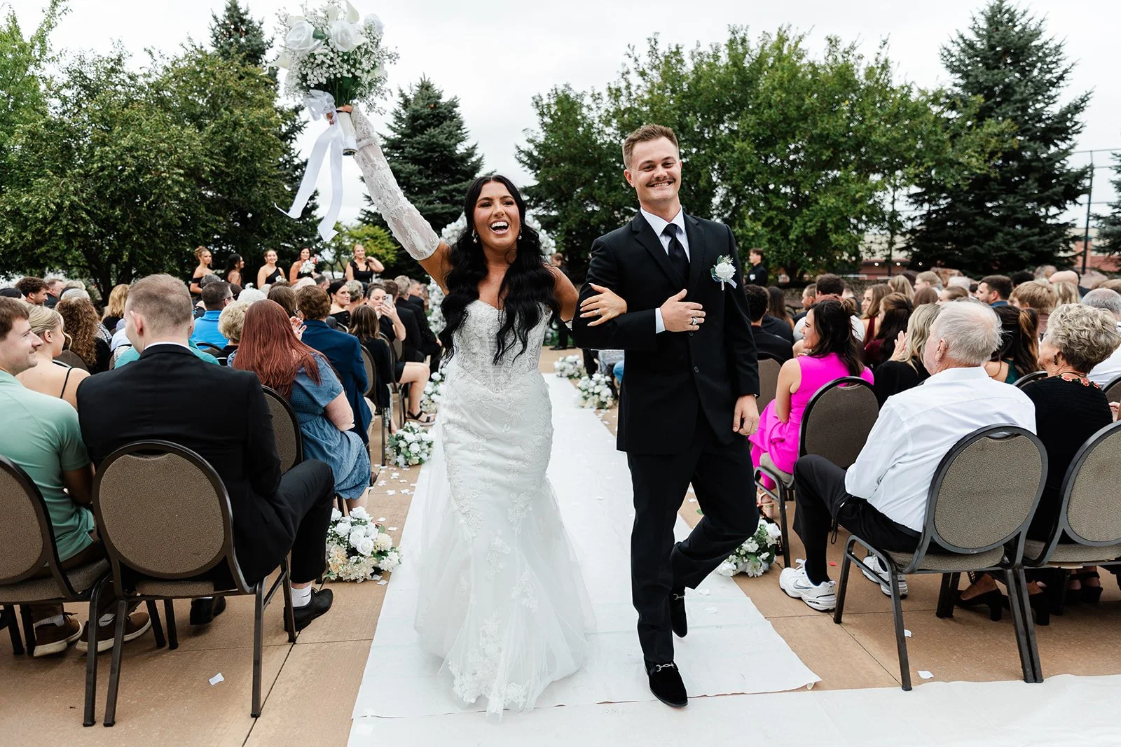 Happy bride and groom walking down the aisle at their outdoor wedding, surrounded by seated guests, with a floral aisle and trees in the background.