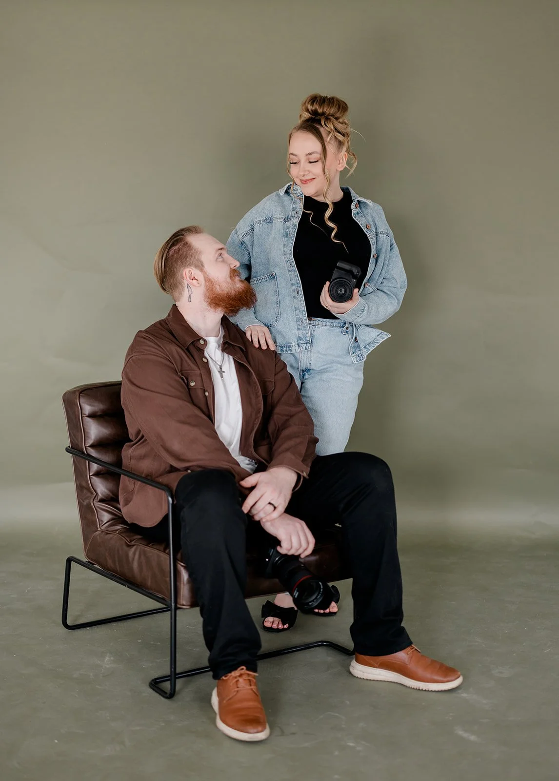 A woman and a man are posing together in a photo studio. The woman is standing, holding a camera, and looking at the man with a smile. The man is sitting on a brown leather chair, looking up at her, holding a camera in his lap. They are both dressed casually, and the background is a plain, neutral color.