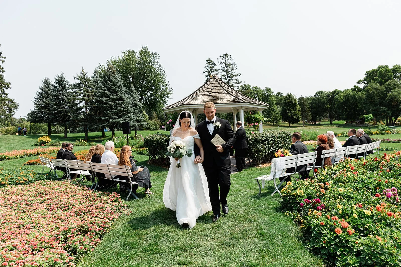 A bride and groom walking down the aisle during their outdoor wedding, surrounded by guests seated on benches in a garden with colorful flowers and trees.