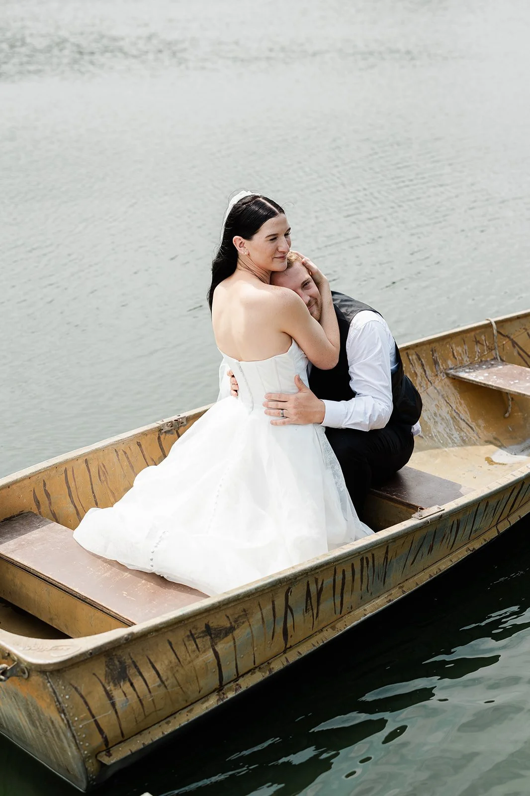 A bride and groom sharing a tender moment on a small boat in a lake, with the bride in a white wedding dress and the groom in a black vest and white shirt.