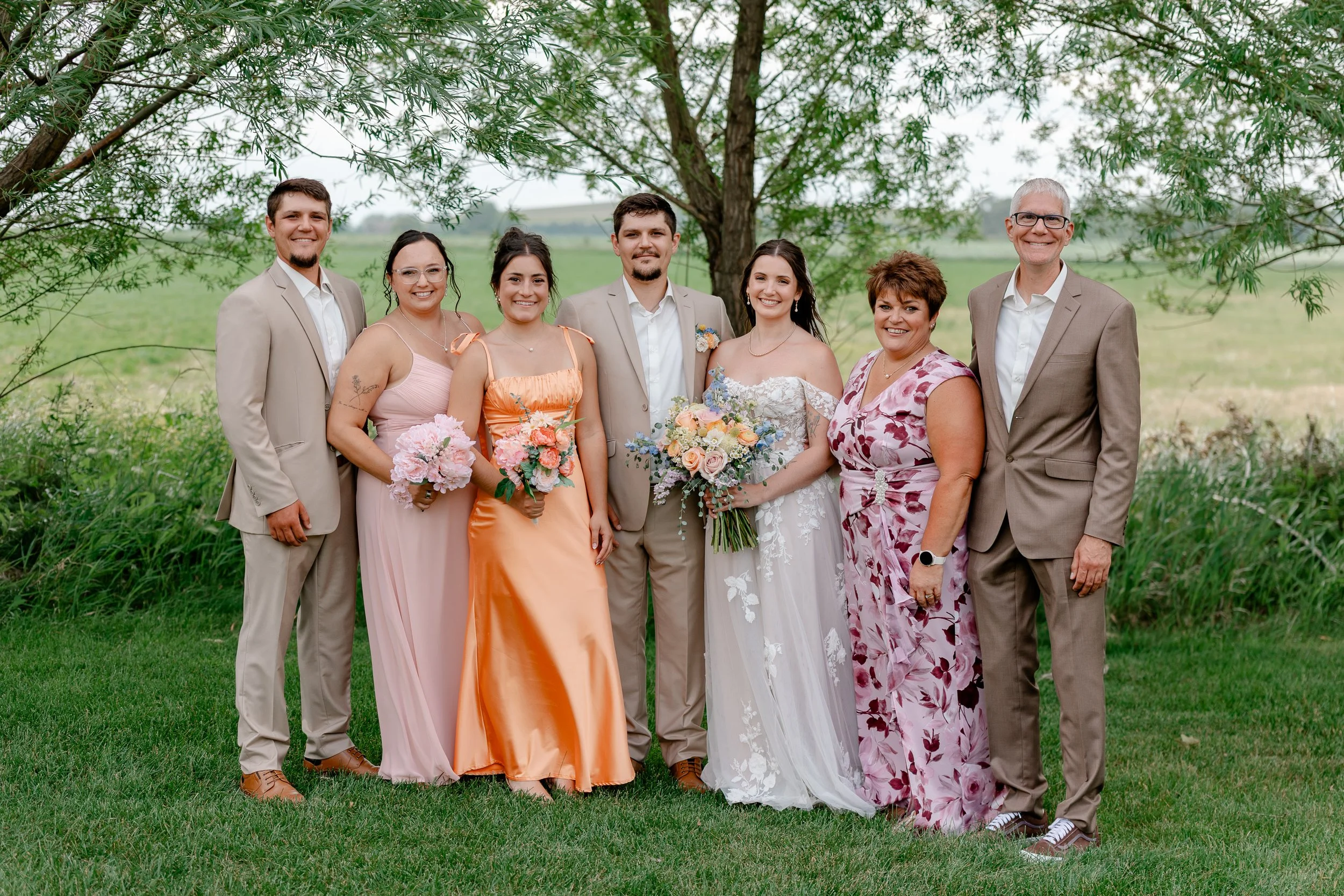 Group of seven people standing outdoors under a tree, dressed in formal attire for a wedding or special event, holding bouquets of flowers, with green fields in the background.