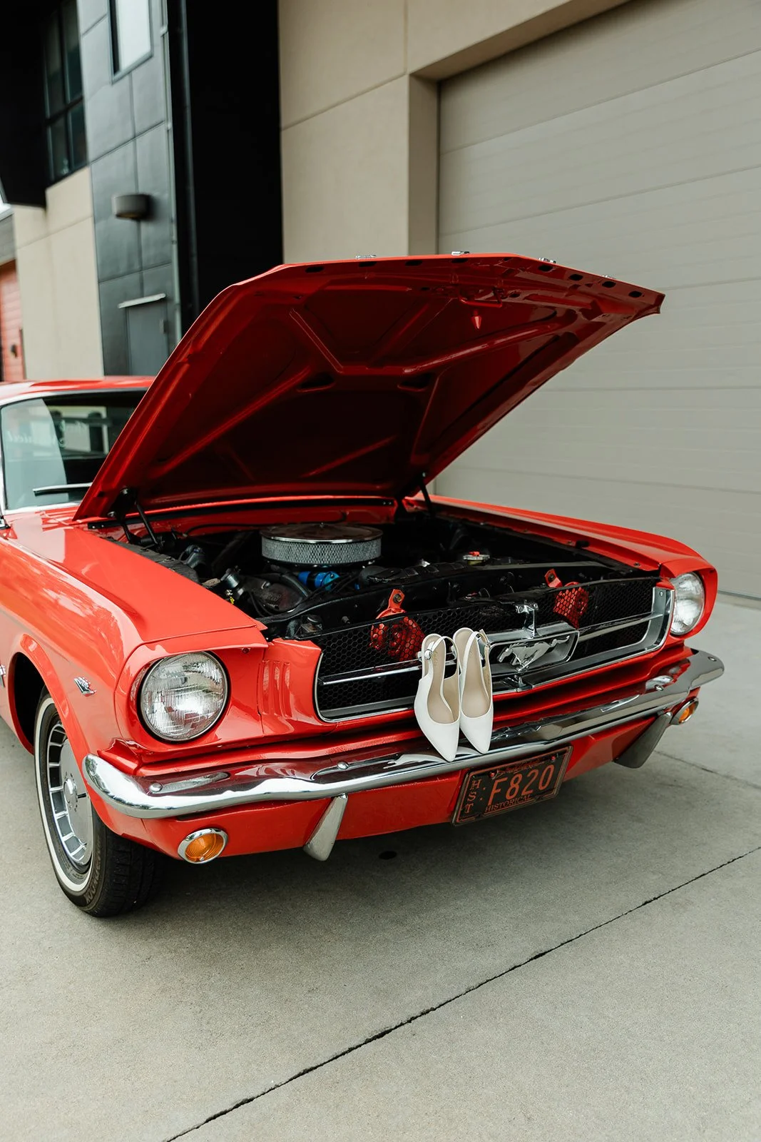 A vintage red car with its hood open, showing the engine, with a pair of white high-heeled shoes hanging from the front grille, parked outside near a modern building.