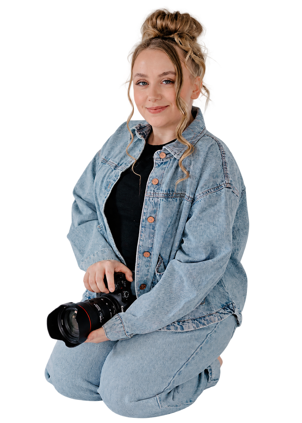 Young woman with blond curly hair styled in a messy bun, holding a professional camera, wearing a blue denim jacket over a black shirt, kneeling with one knee up, smiling at the camera.
