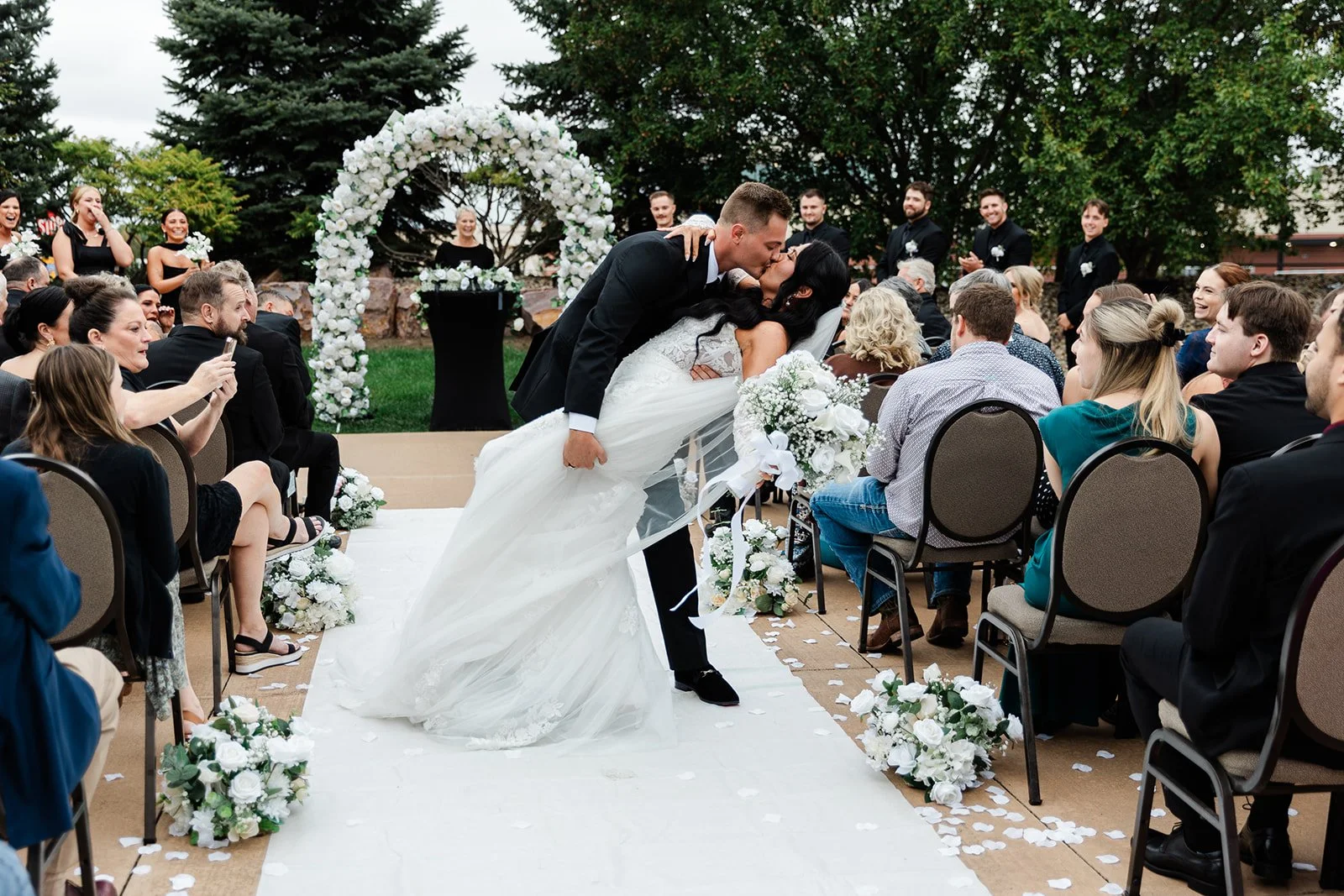 A newlywed couple sharing a kiss during an outdoor wedding ceremony, with guests seated and smiling around them, decorated with white flowers and greenery.
