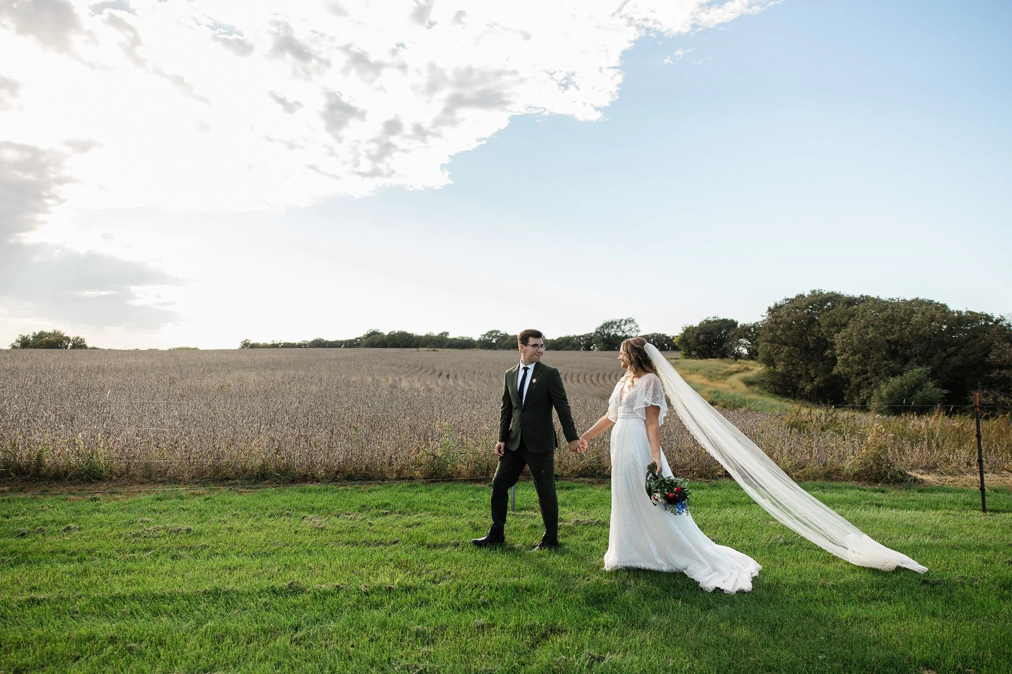 A bride and groom holding hands and walking outdoors in a field during their wedding, with a cloudy sky overhead and trees in the background.