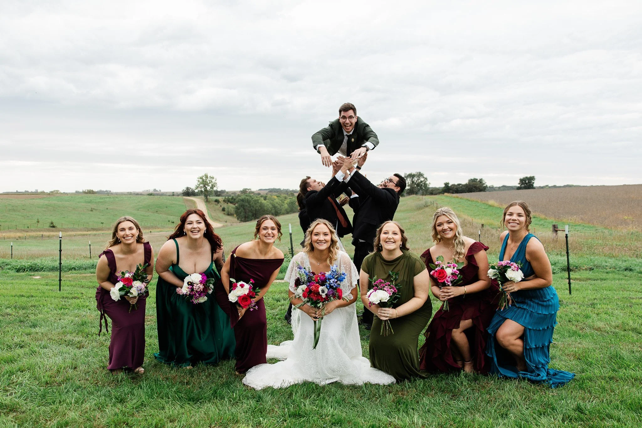 Group of women dressed in colorful bridesmaid dresses and a bride in white gown holding bouquets, standing on grass outdoors with a group of men lifting a man in suit above them, under cloudy sky.