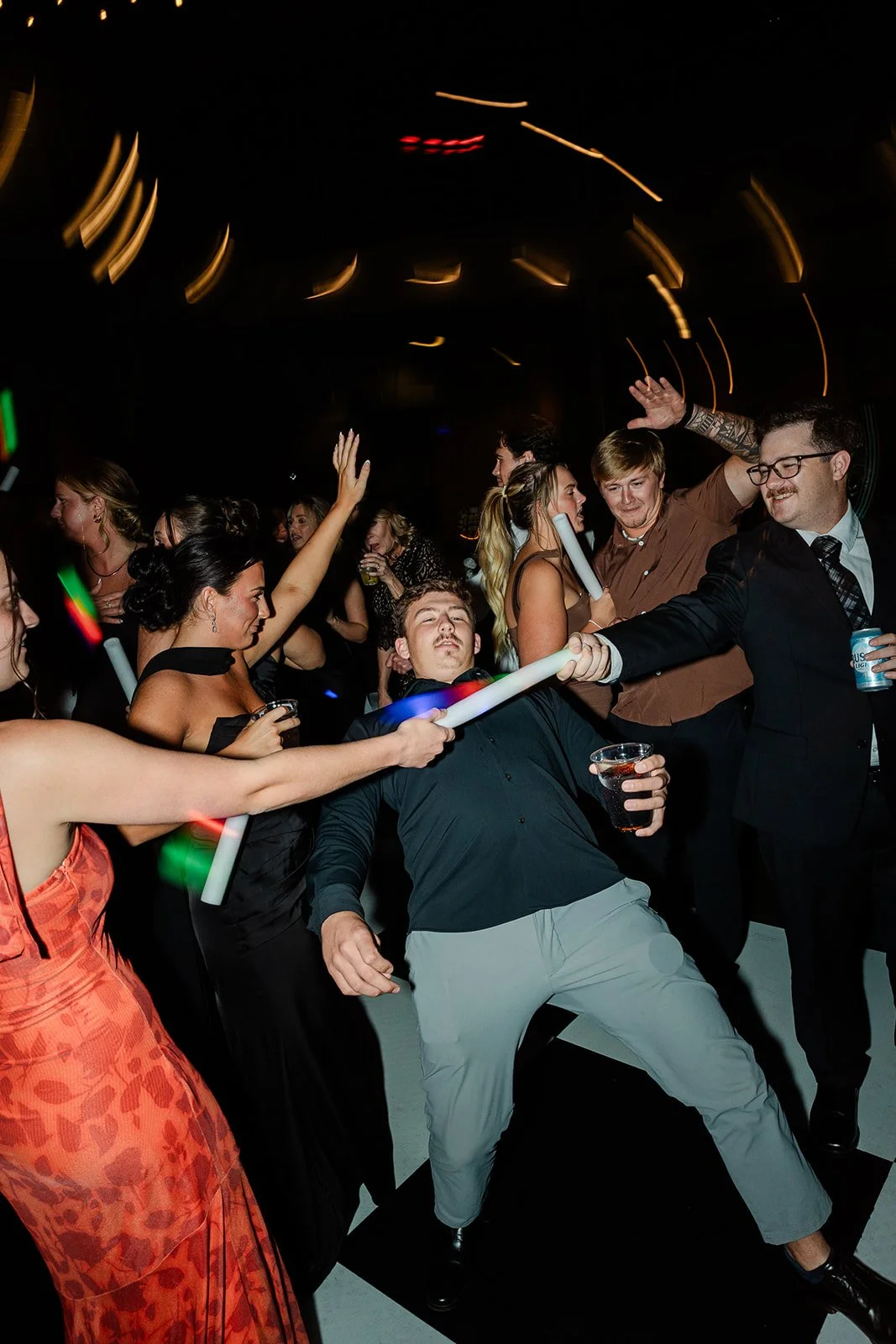 People dancing and celebrating at a party, with some holding drinks and glow sticks, in a dark room with blurred lights.