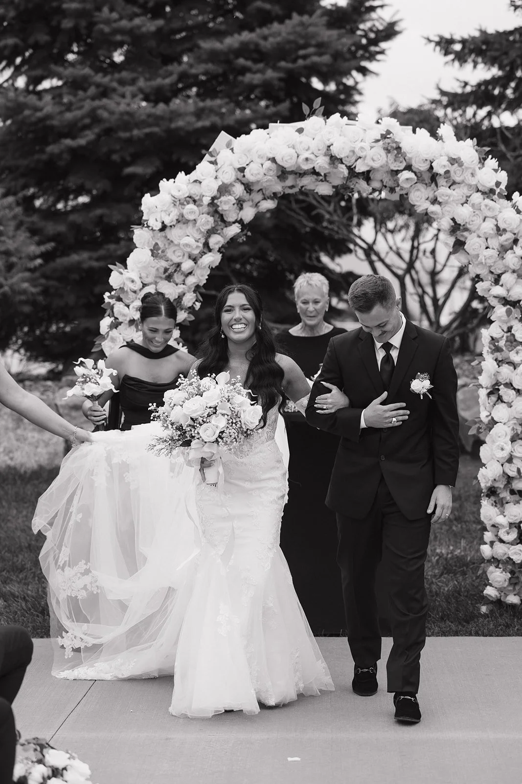 A black and white photo of a bride and groom walking under a floral arch, with bridesmaids and an officiant celebrating.