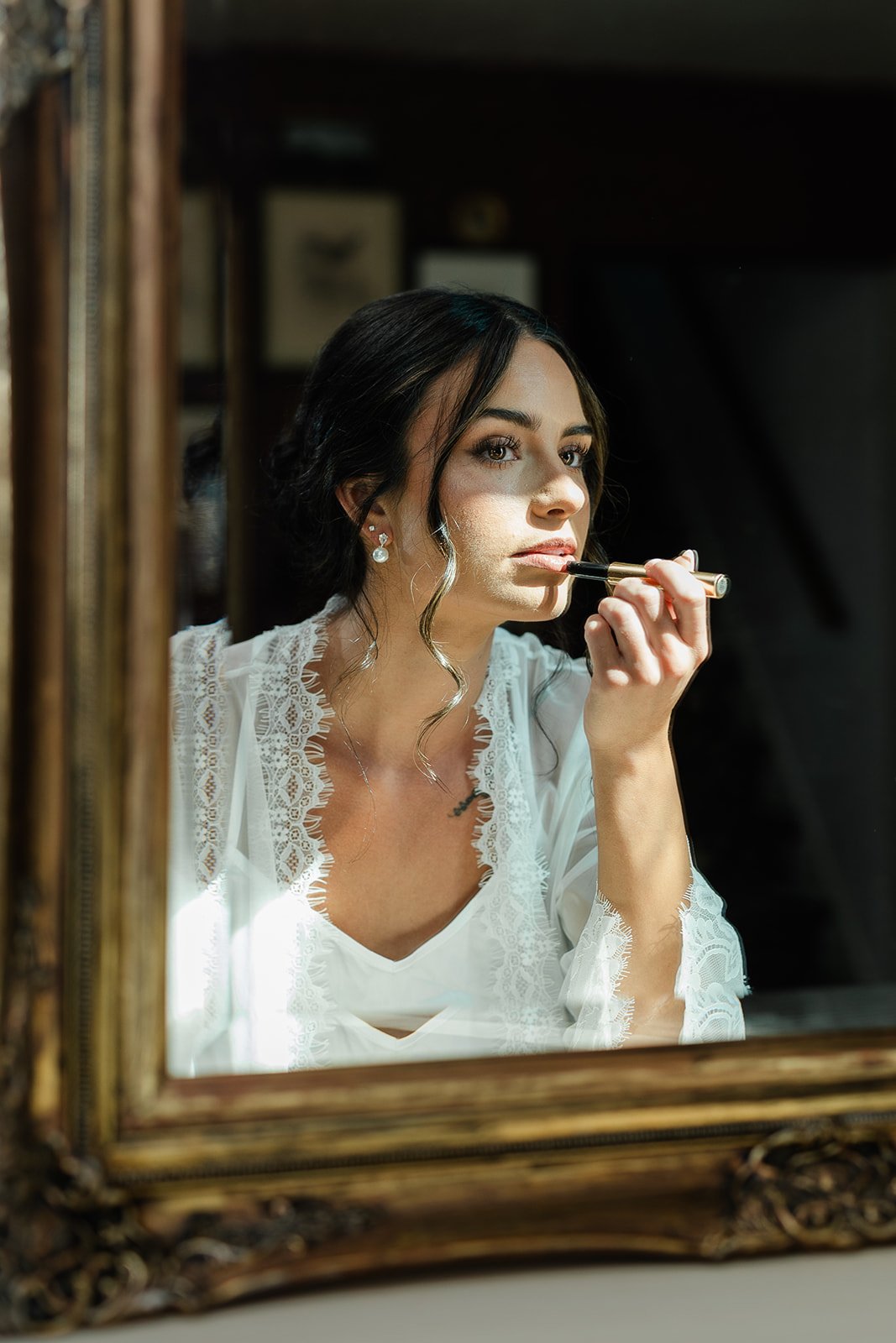 A woman with dark hair and earrings is applying lipstick while looking in a mirror.