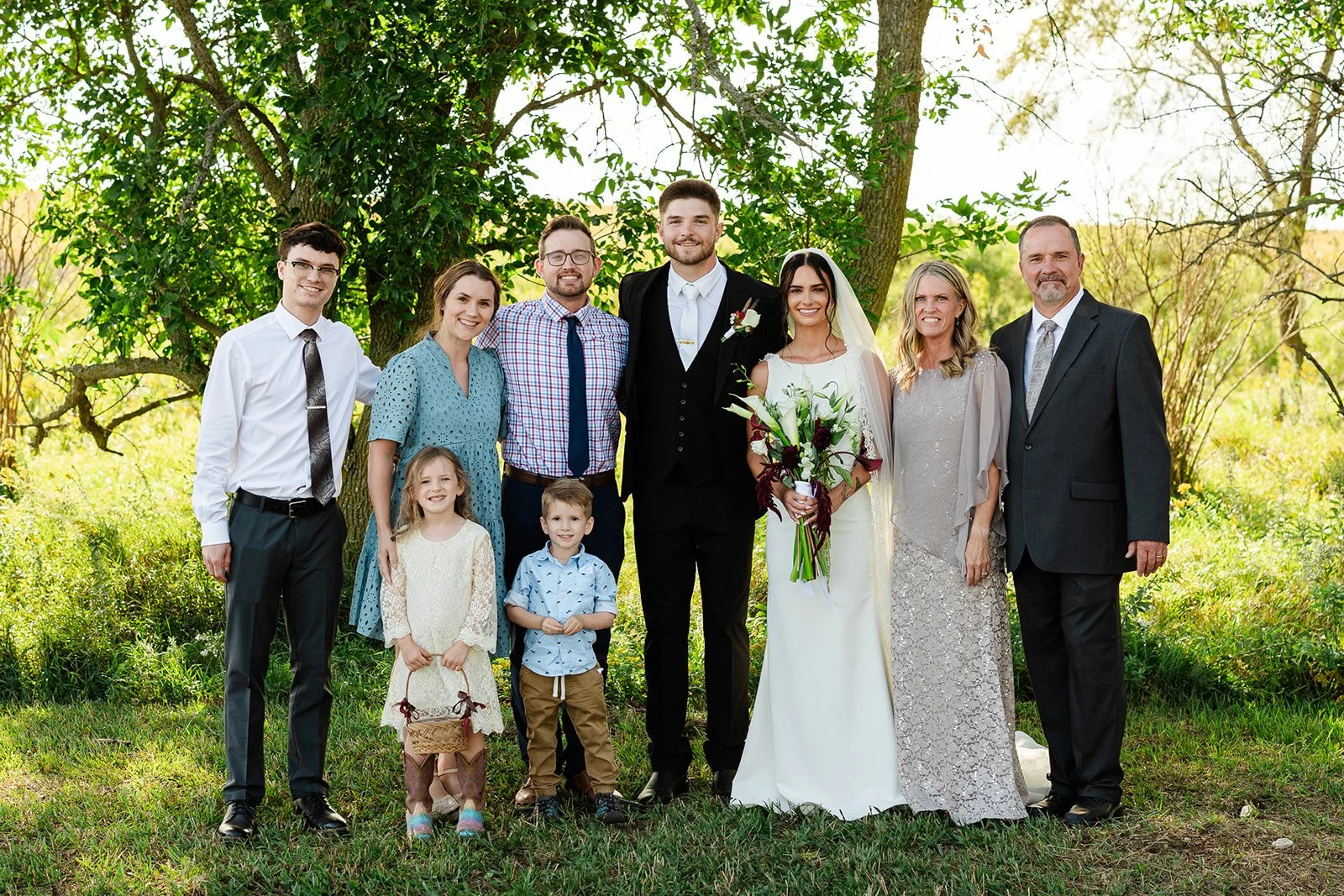 A wedding photo featuring a bride and groom with family members outdoors under a tree, with greenery and sunlight in the background.