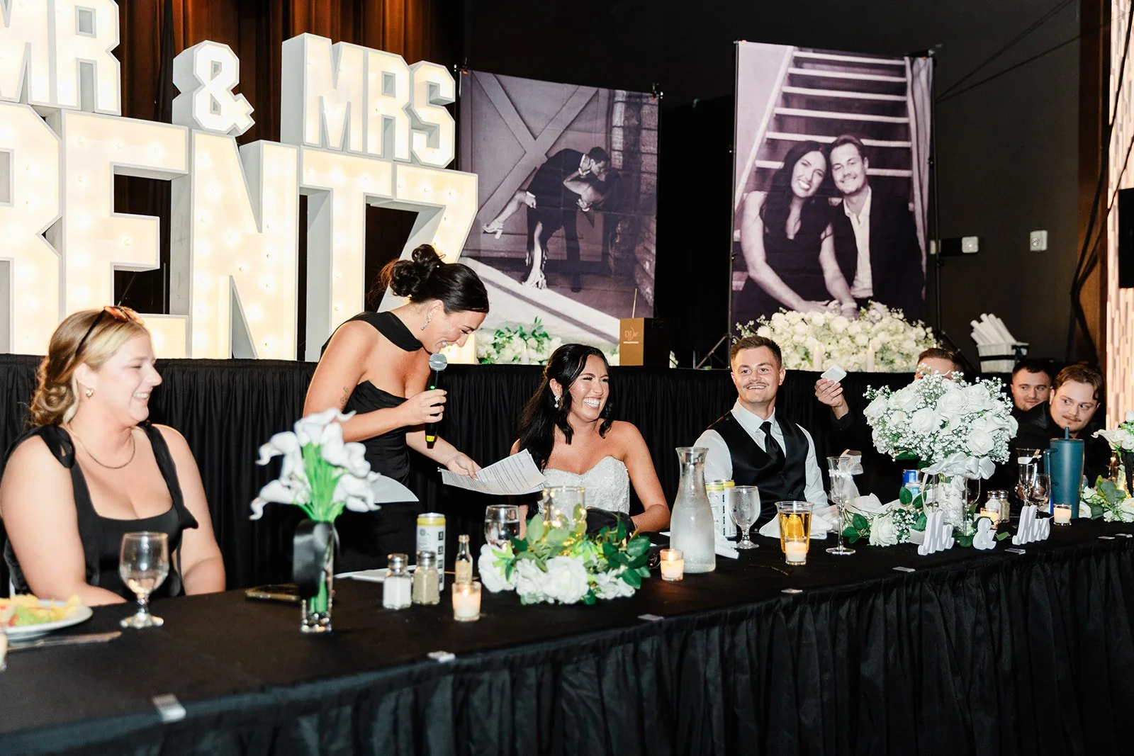 A wedding reception scene with the bride and groom sitting at a decorated table, surrounded by bridesmaids and friends. The bride and groom are smiling, with the bride in a white dress and the groom in a black vest and white shirt. A woman in a black