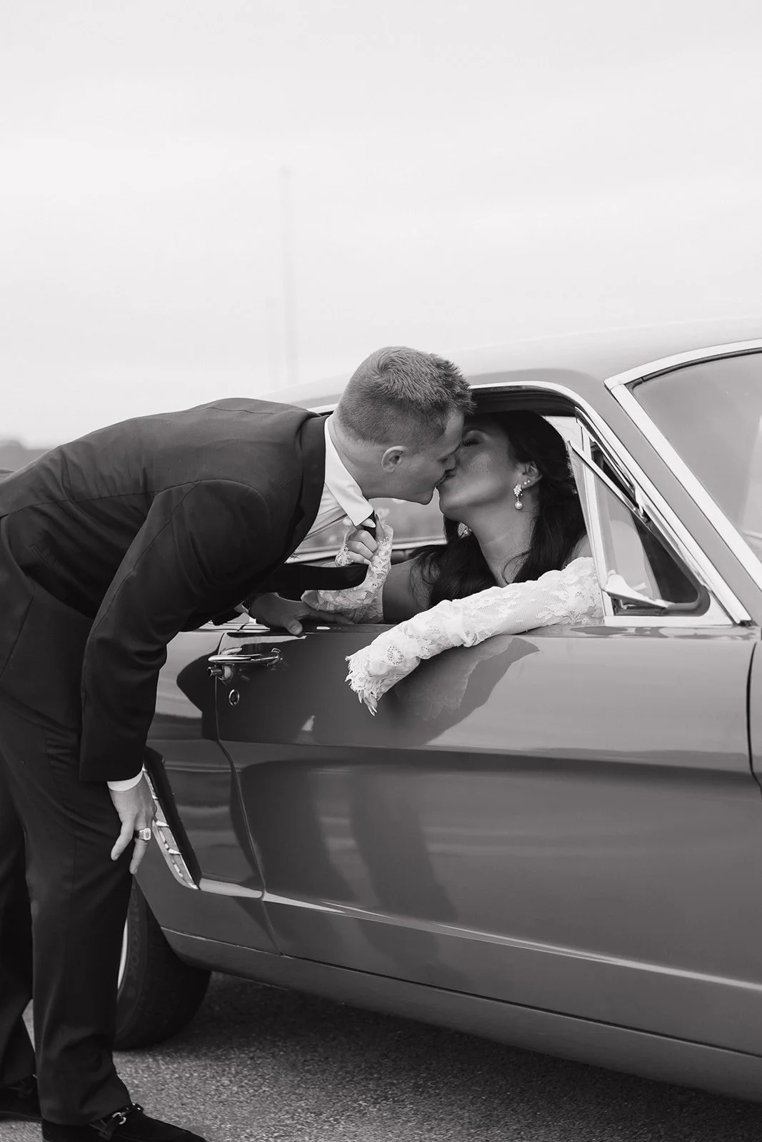 A black-and-white photo of a man in a suit leaning into a car window to kiss a woman in a wedding gown, on a cloudy day.