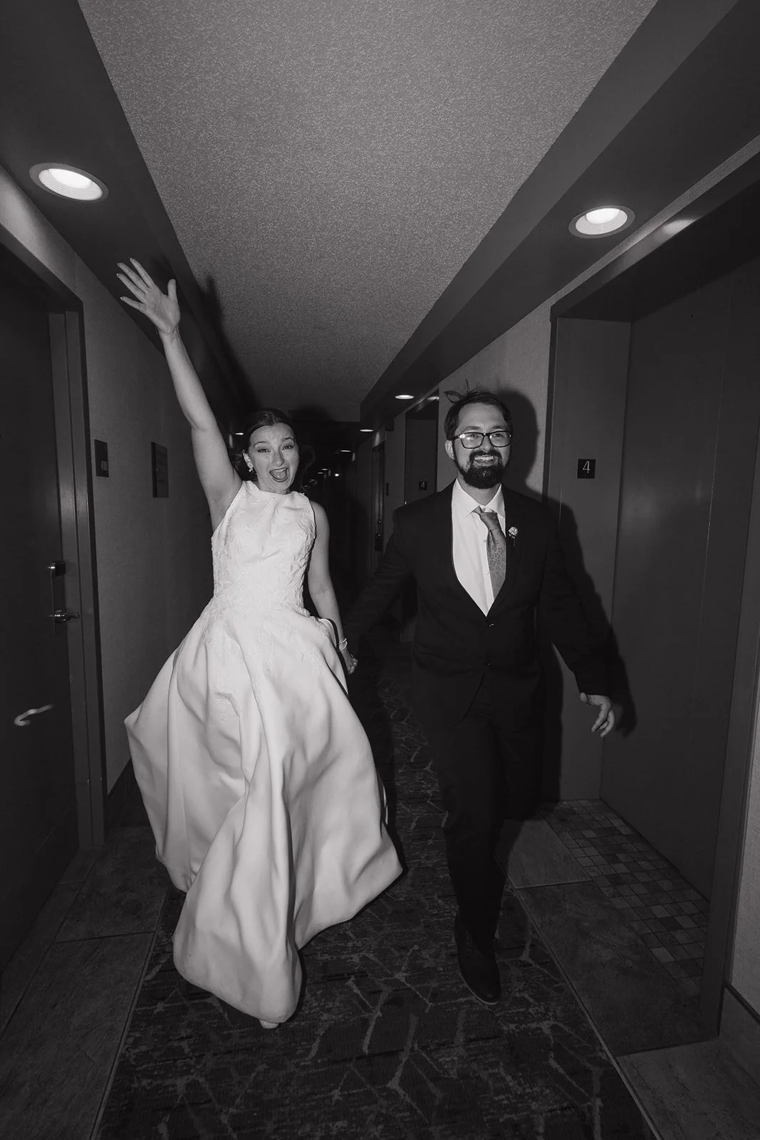 Black and white photo of a joyful bride and groom running down a hotel corridor. The bride in a wedding dress with her arm raised, the groom in a suit with glasses, both smiling.