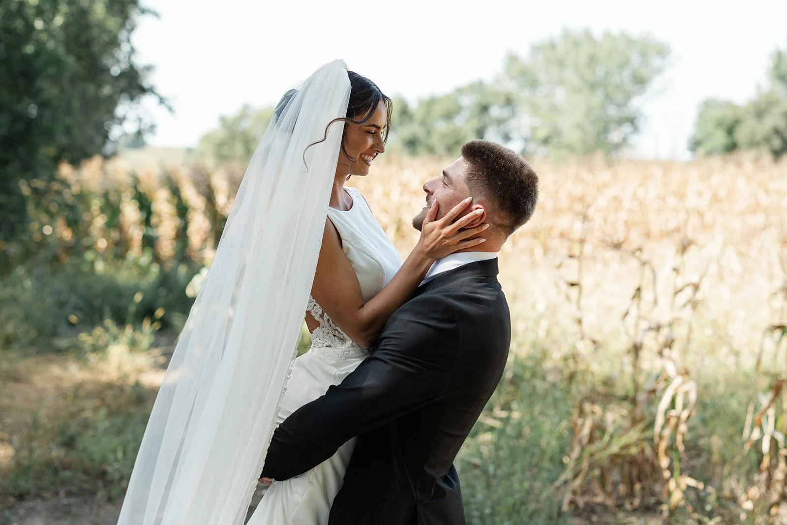 A bride and groom in wedding attire sharing a joyful moment outdoors, with the groom lifting the bride. The bride is smiling, wearing a white wedding gown and veil, while the groom is in a black tuxedo. The background includes trees and bushes.