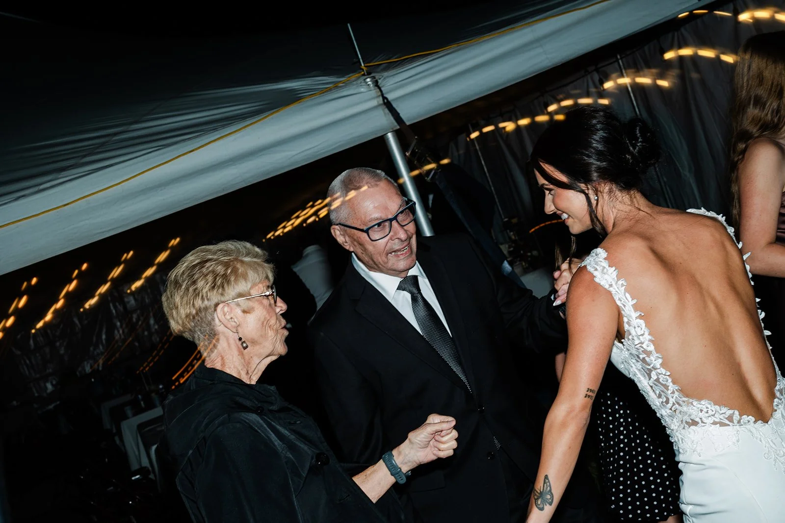 People at a wedding celebration, with a woman in a white dress, talking to an older woman and an older man, under a decorated tent with hanging lights.